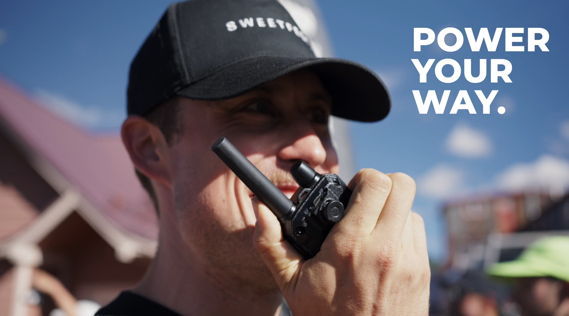 A man wearing a black cap outdoors speaking into a walkie-talkie with the text 'Power Your Way.' in white on the right side.