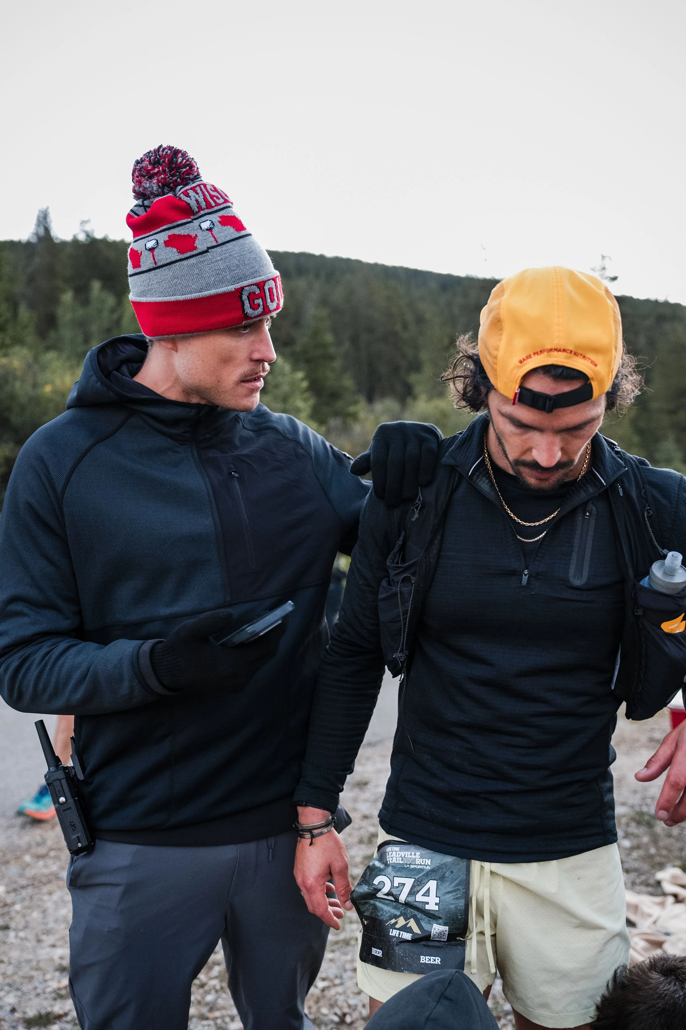 Two men, one with a red and gray beanie and the other with a yellow cap, are outdoors. One is holding a phone and has a walkie-talkie attached to his pants. They appear to be in a race, as indicated by the bib number 274 on the shorts of the man with the yellow cap. The man with the yellow cap is wearing a black jacket and beige shorts, and the man with the beanie has his arm around him.