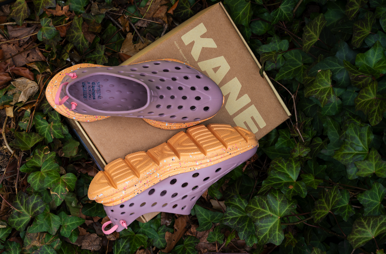 Pink and yellow children's shoes with holes, placed on top of a cardboard shoe box labeled 'KANDY', surrounded by green ivy leaves and brown fallen leaves.