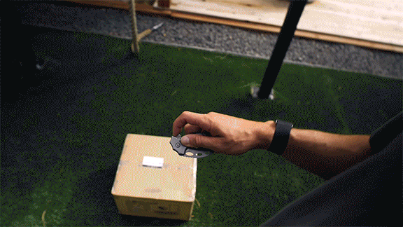 A person holding a small gray folding knife above a cardboard box outdoors.