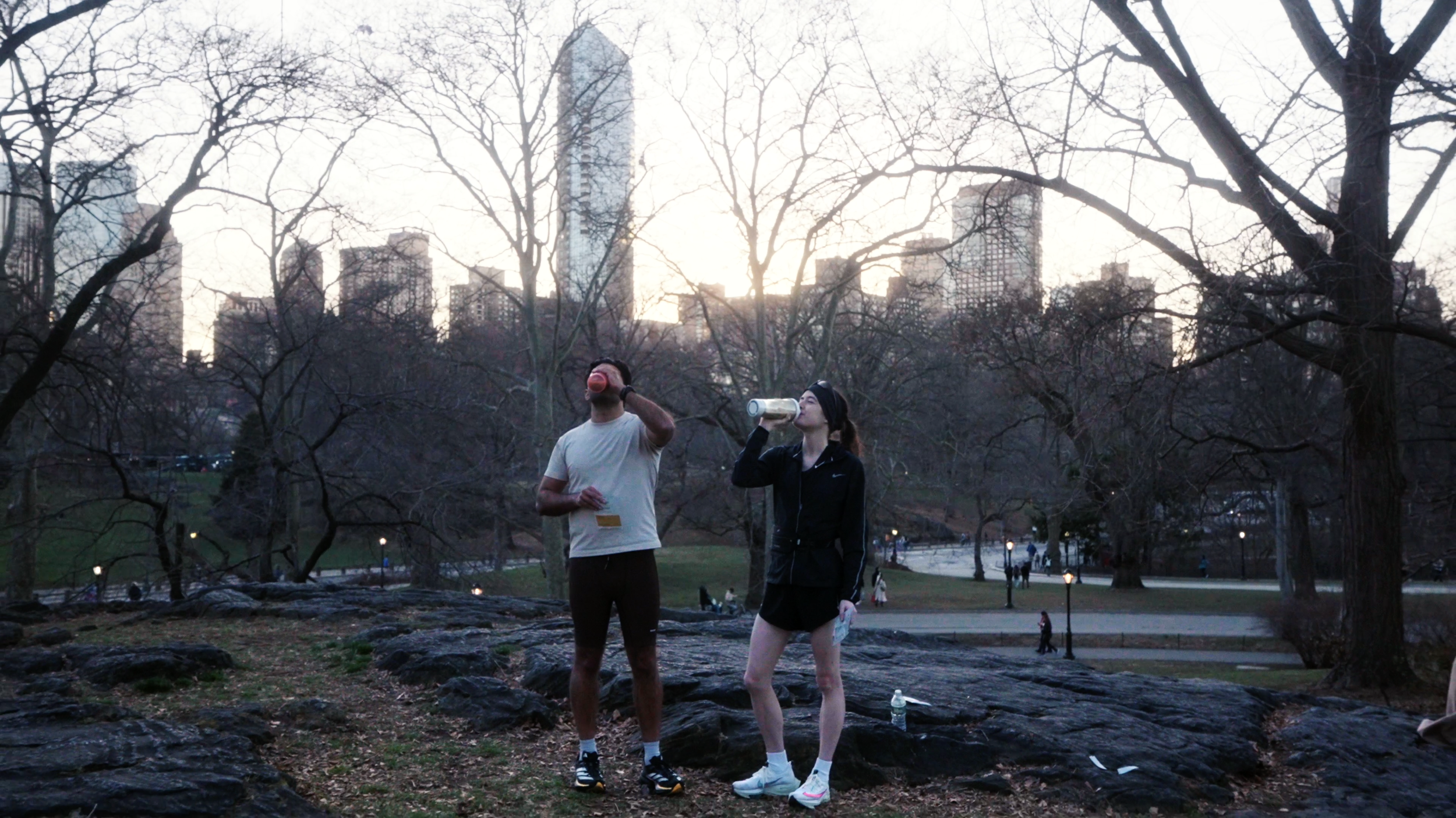 Two people in athletic clothing drinking water from bottles in a park during dusk, with city skyscrapers in the background and bare trees around.