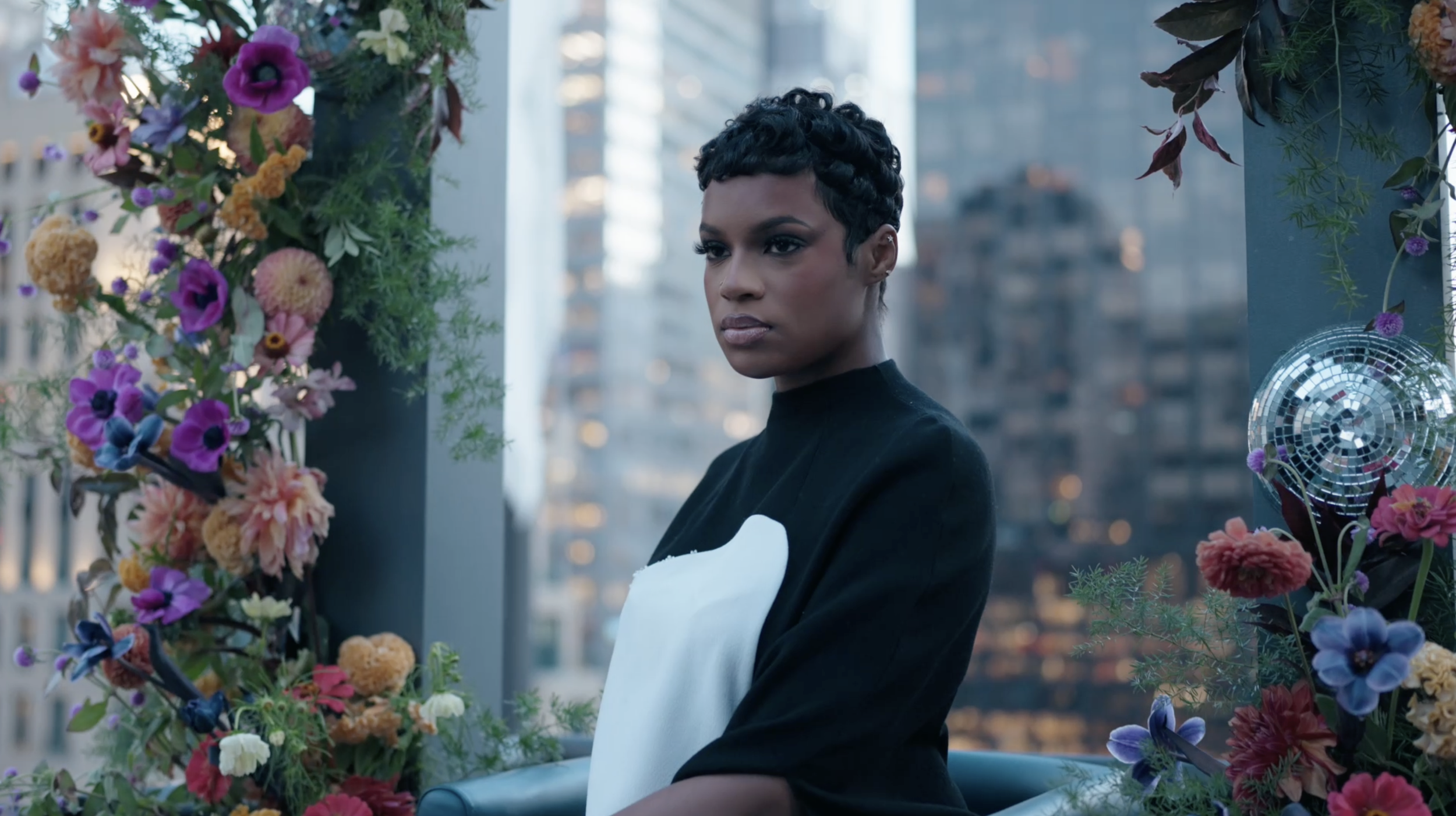 A woman with short curly hair and dark skin, wearing a black and white top, posing between colorful flowers with a cityscape in the background.