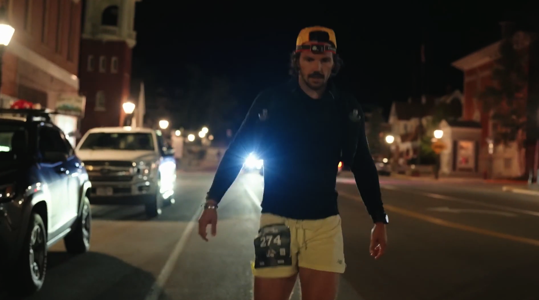 A man with a headlamp on his forehead, wearing a black long-sleeve shirt and yellow shorts, walking on a street at night with parked cars and buildings in the background.
