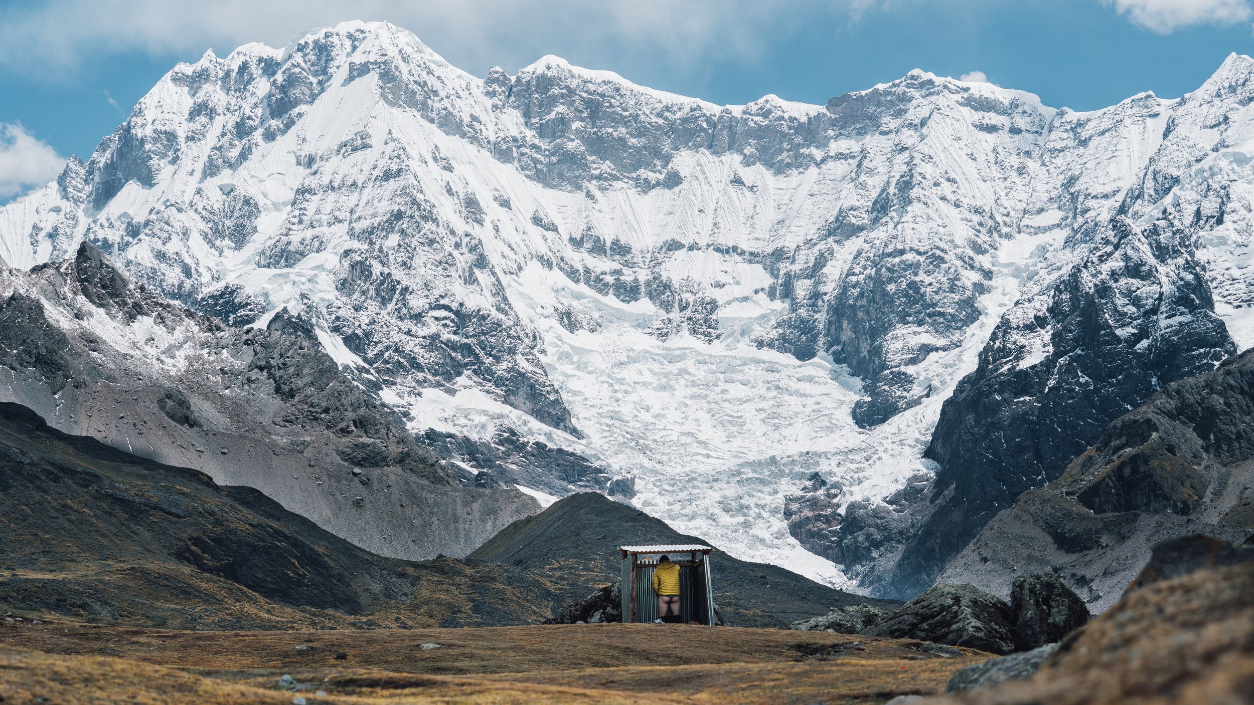 A person standing at a small shelter in a valley with snow-capped mountains in the background.