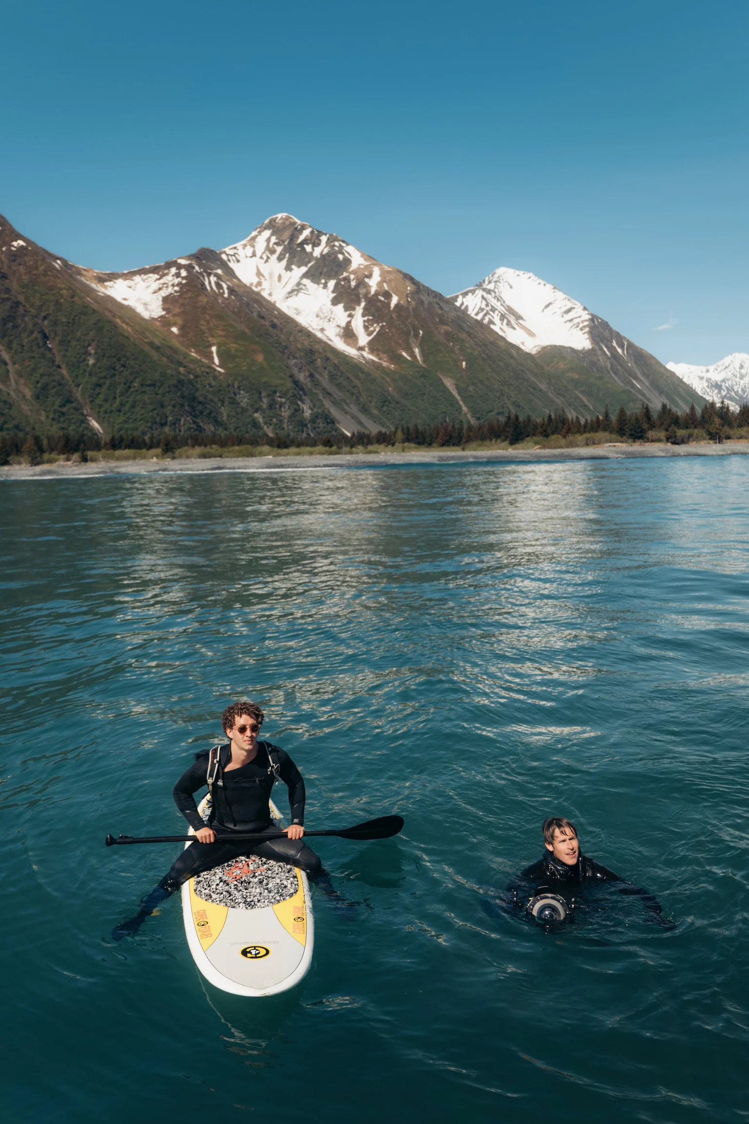 A person paddleboarding on a lake with mountains in the background, and another person swimming nearby.