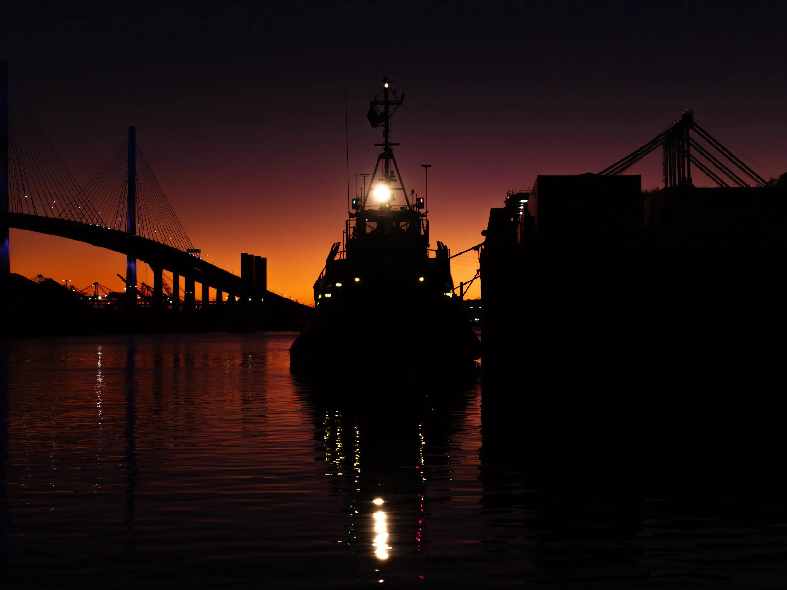 Silhouette of a boat docked at a harbor during sunset, with a bridge in the background and colorful sky