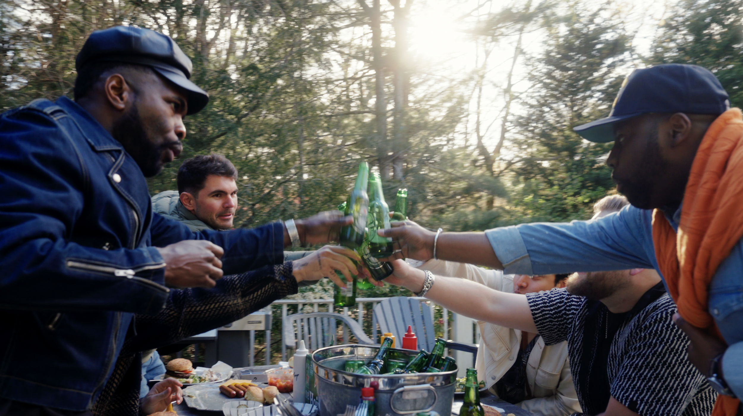 A group of people sharing drinks outdoors, clinking green beer bottles during a gathering in a wooded area with sunlight filtering through trees.