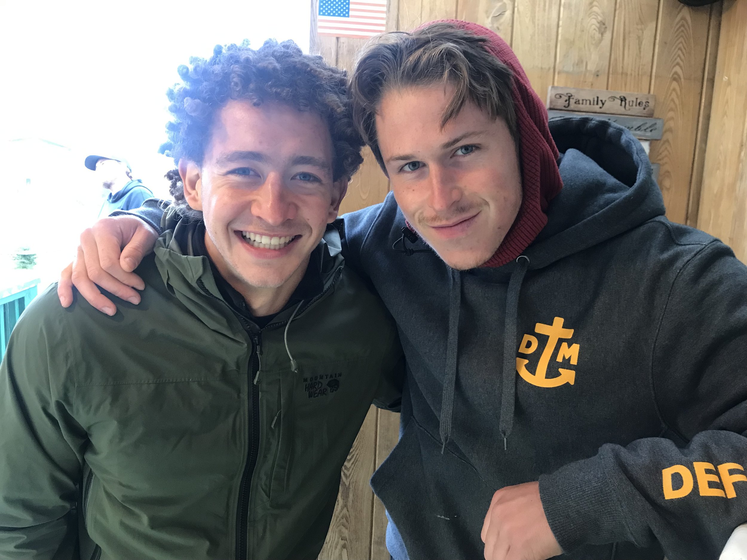 Two young men smiling, with one having curly hair and wearing a green jacket, and the other with straight hair and wearing a dark hoodie with a yellow logo, inside a wooden-paneled room.