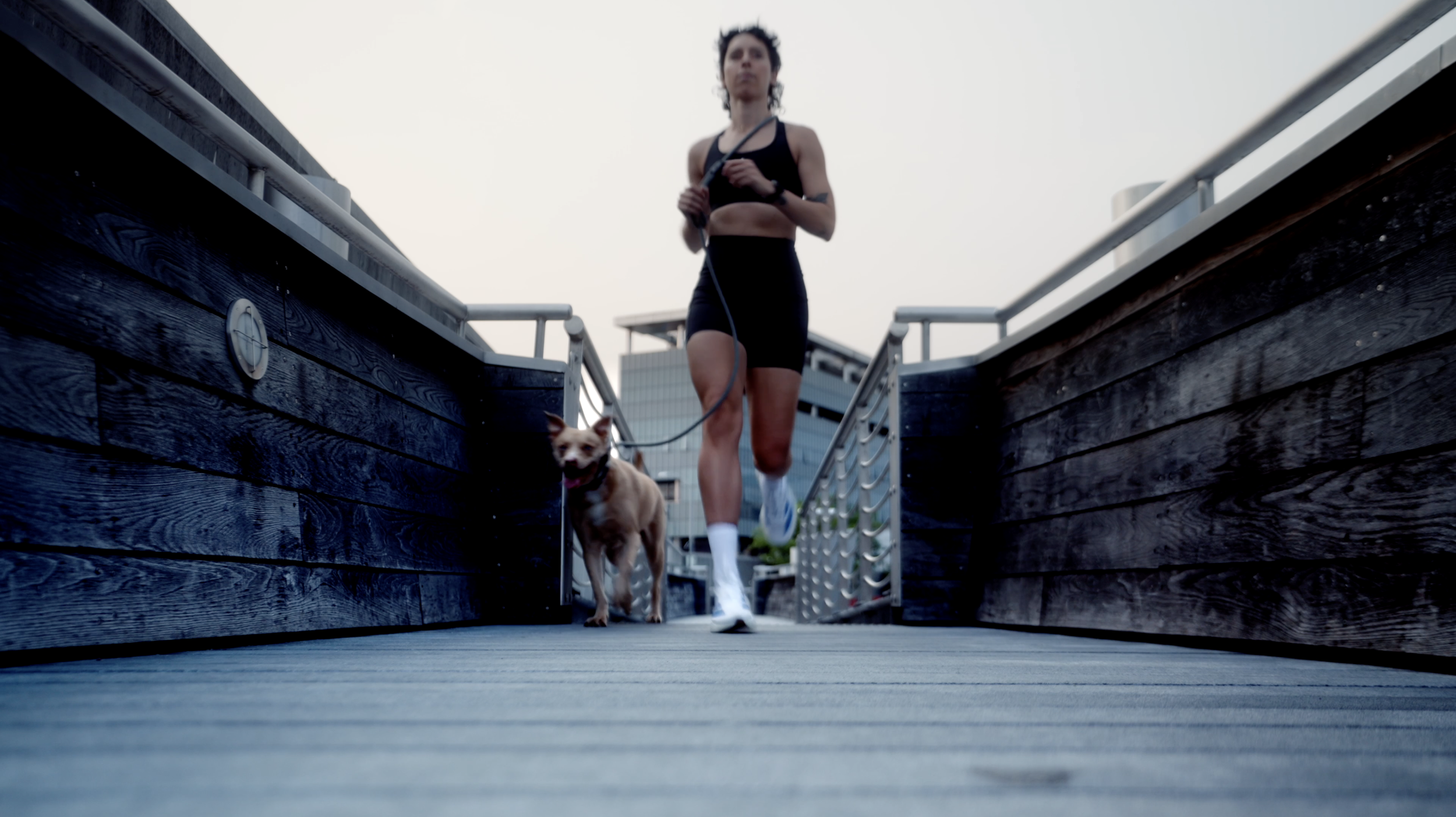 A woman running on a wooden pier with her dog on a leash, with urban buildings in the background.