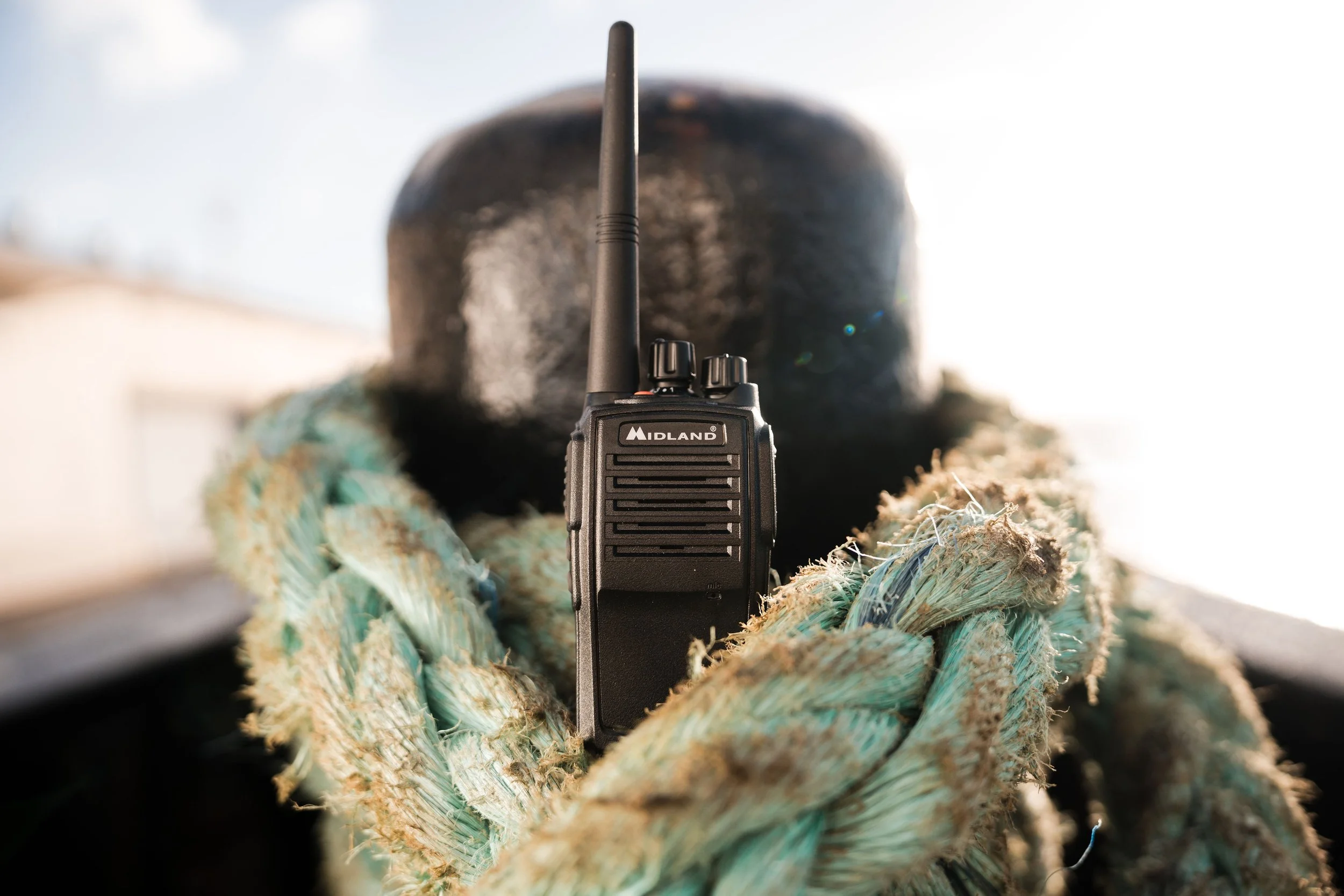 Black Midland walkie-talkie radio held by a person wearing a camouflage knit glove, with a black hat in the background and bright sunlight.