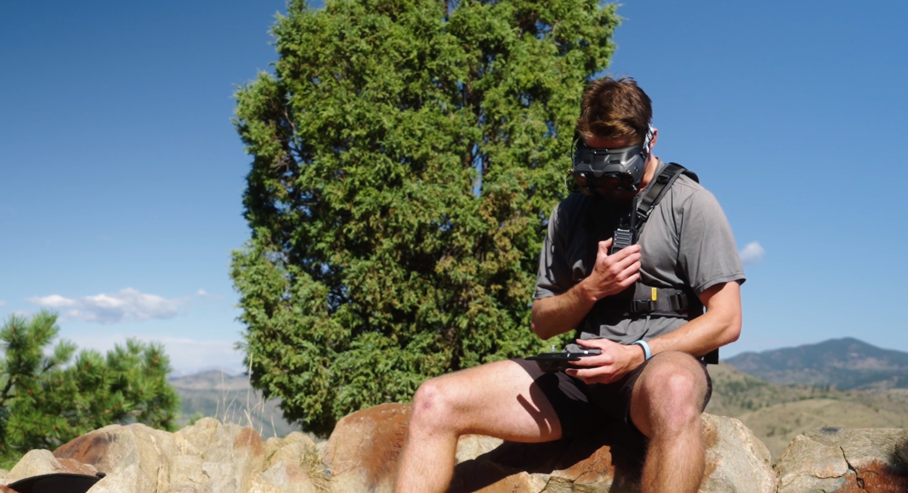 A man in outdoor gear sits on a large rock in a natural landscape, wearing a virtual reality headset and holding a controller, with trees and mountains in the background.