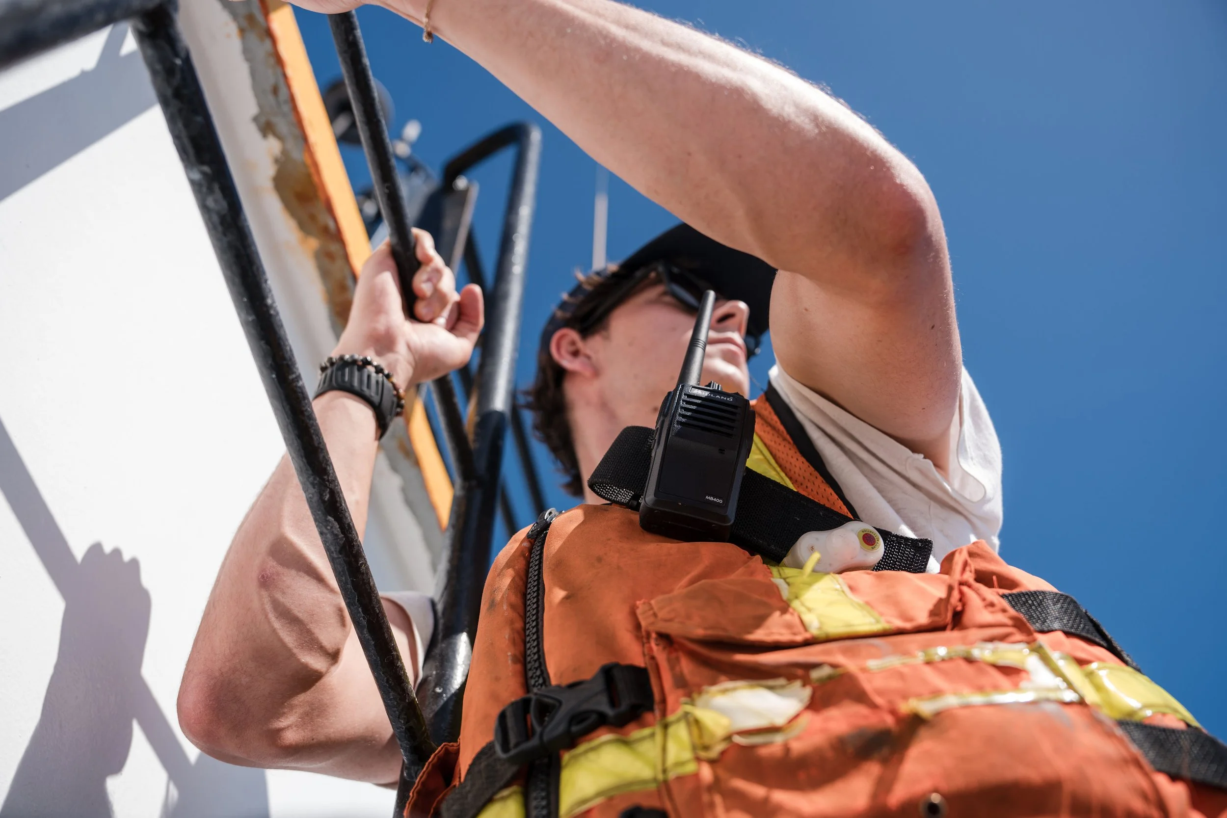 A rescue worker in an orange safety vest and helmet is on a ladder, using a hand to signal or hold onto the ladder, with a communication device attached to their vest against a bright blue sky.