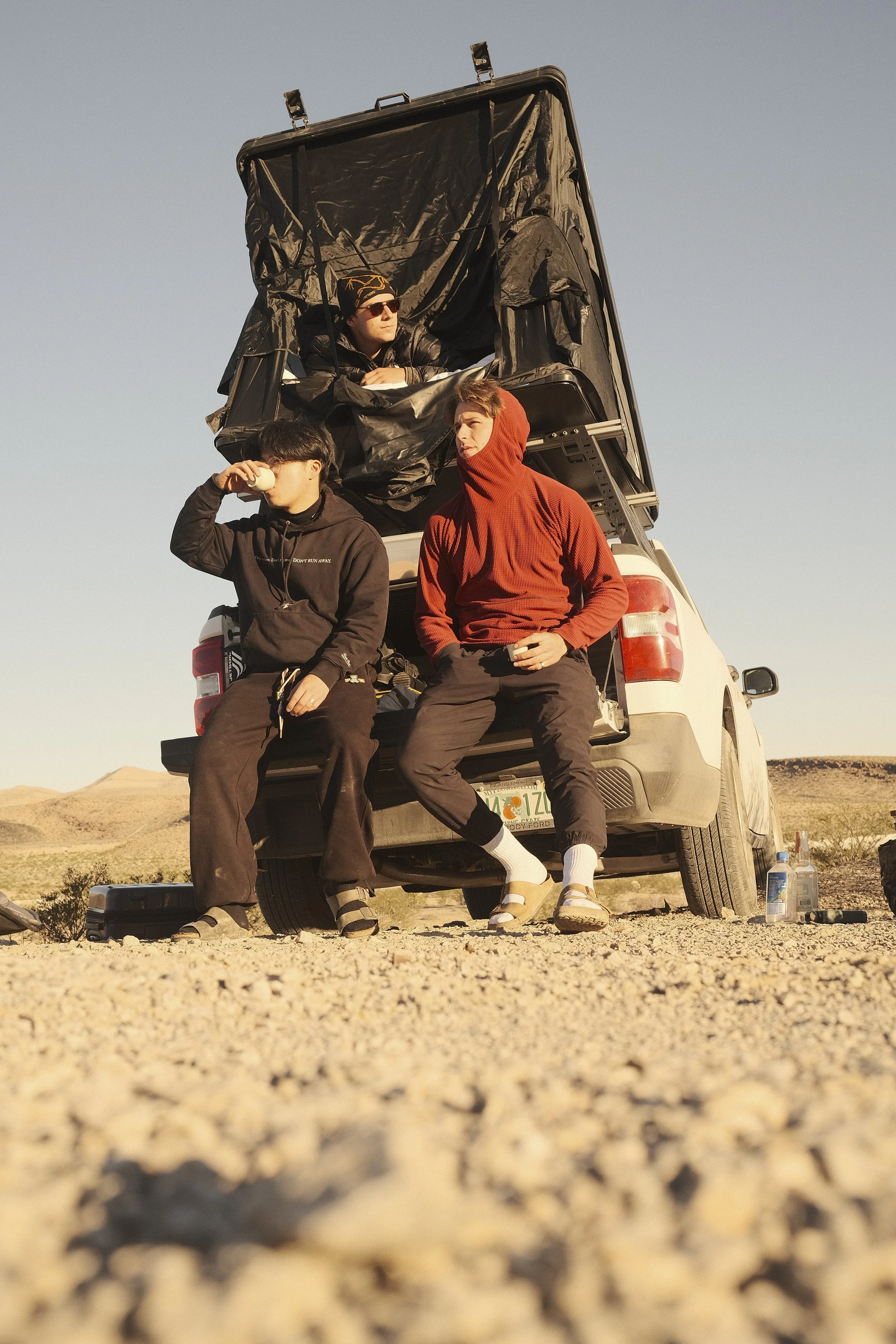 Three young men are relaxing by their vehicle in a desert landscape with sand and hills in the background. One is sitting on the back of the vehicle drinking from a cup, another is sitting on the bumper with a hood over his head, and the third is sit