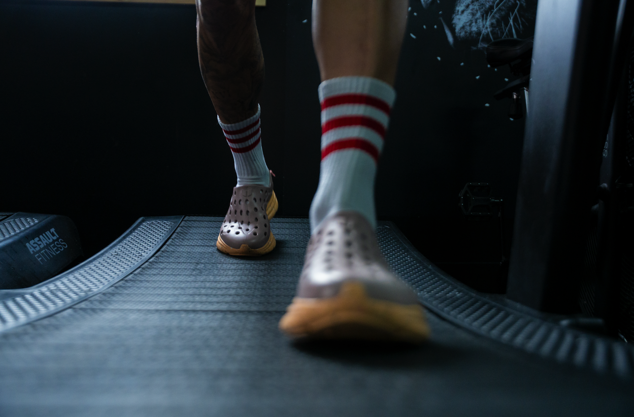 Person walking on a treadmill wearing striped socks and Crocs shoes with a tattooed leg visible.