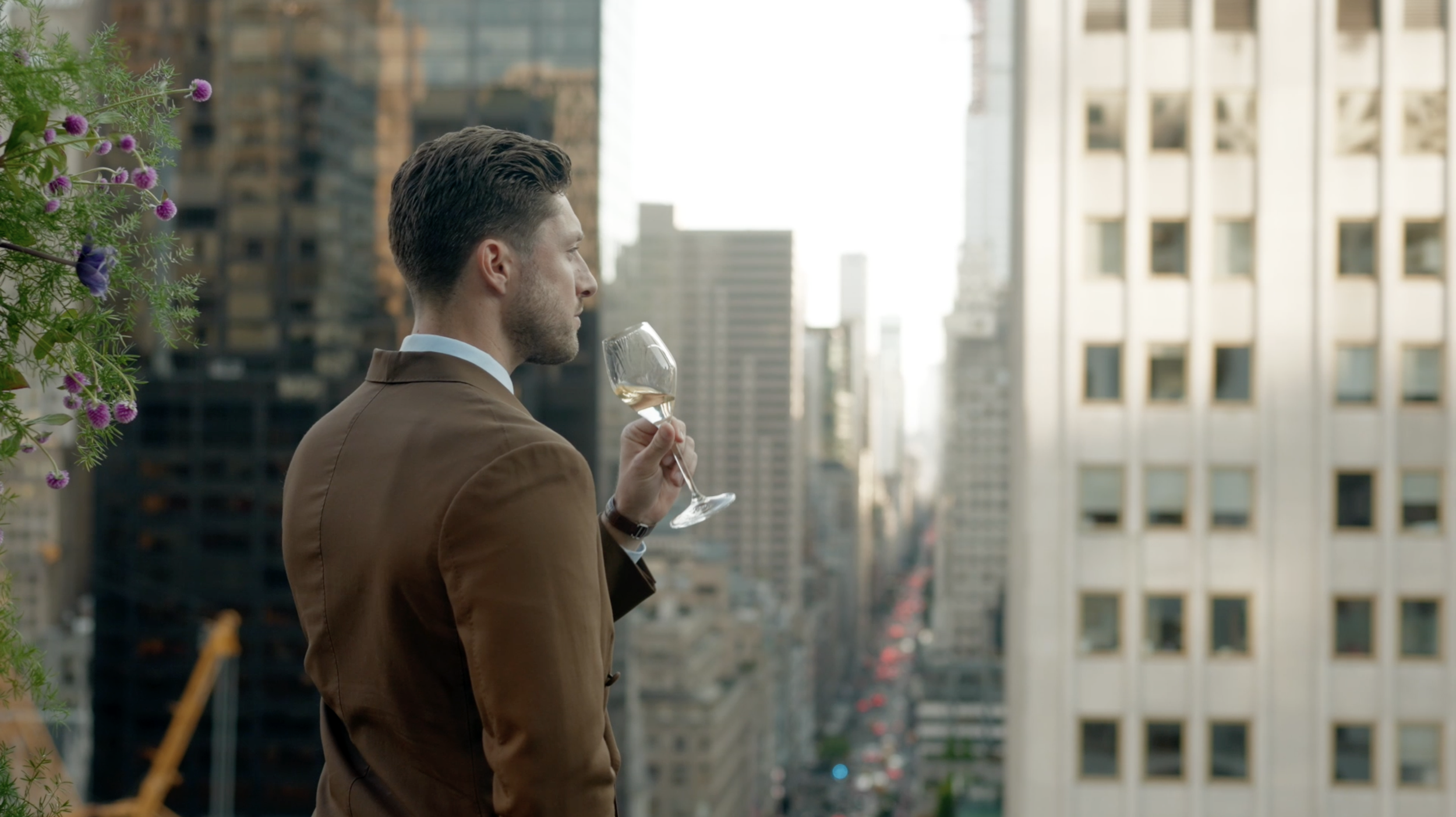 A man in a brown suit with a white shirt holding a glass of white wine on a balcony overlooking a city with tall buildings.