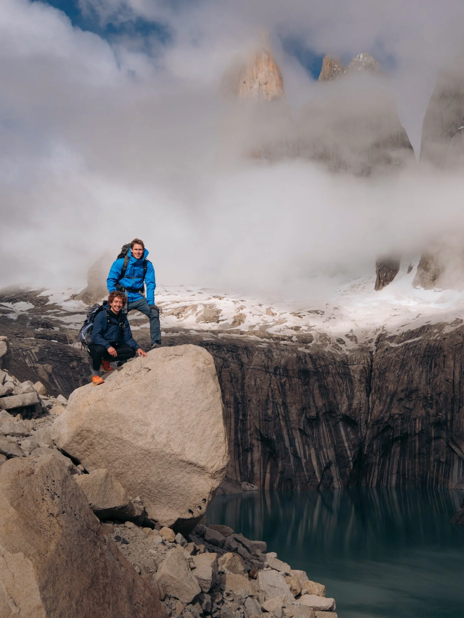 Two hikers in blue jackets and backpacks pose on rocks near a mountain lake surrounded by rocky terrain and snow, with misty clouds and mountain peaks in the background.