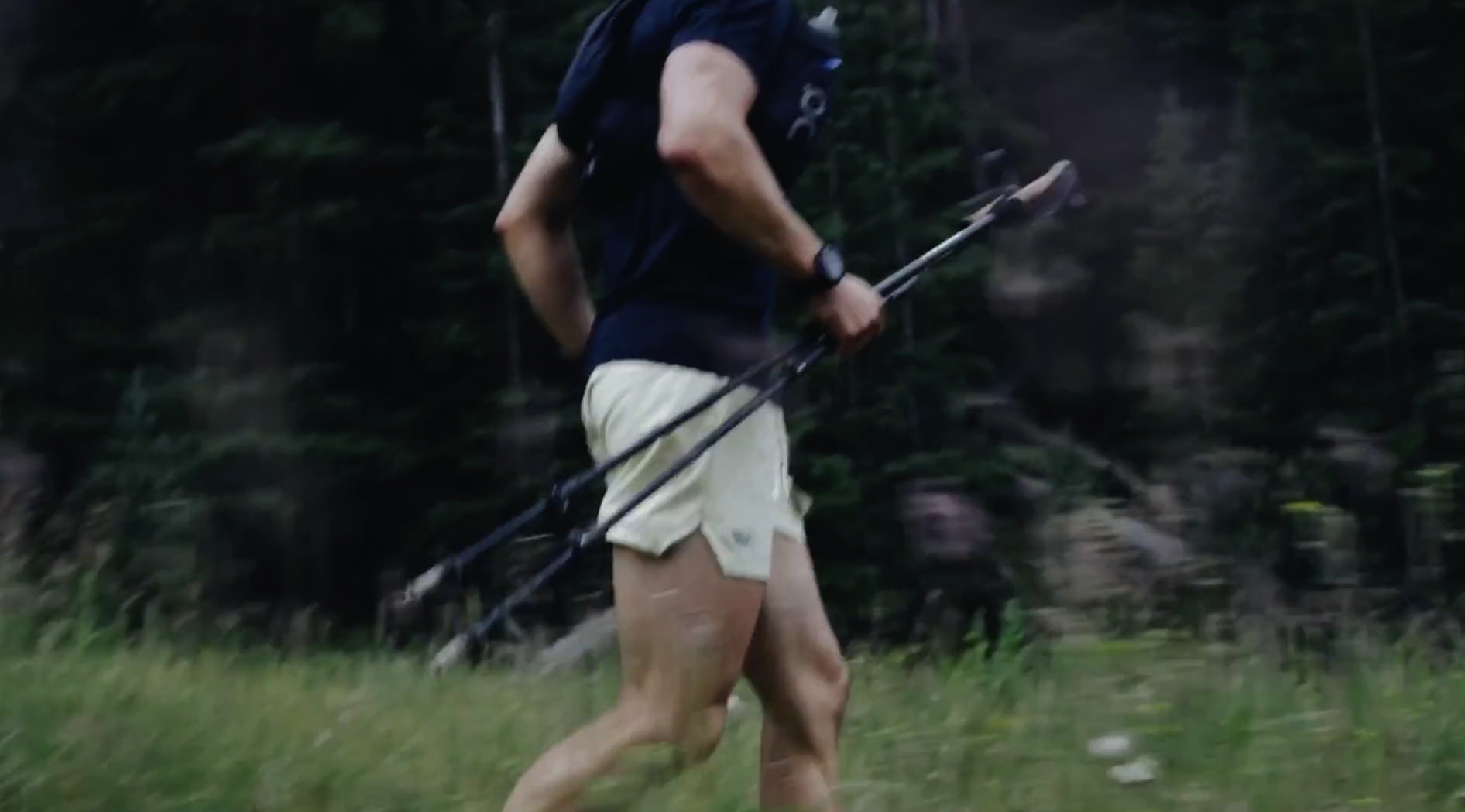A man running outdoors on grass, holding trekking poles, with dense trees in the background.