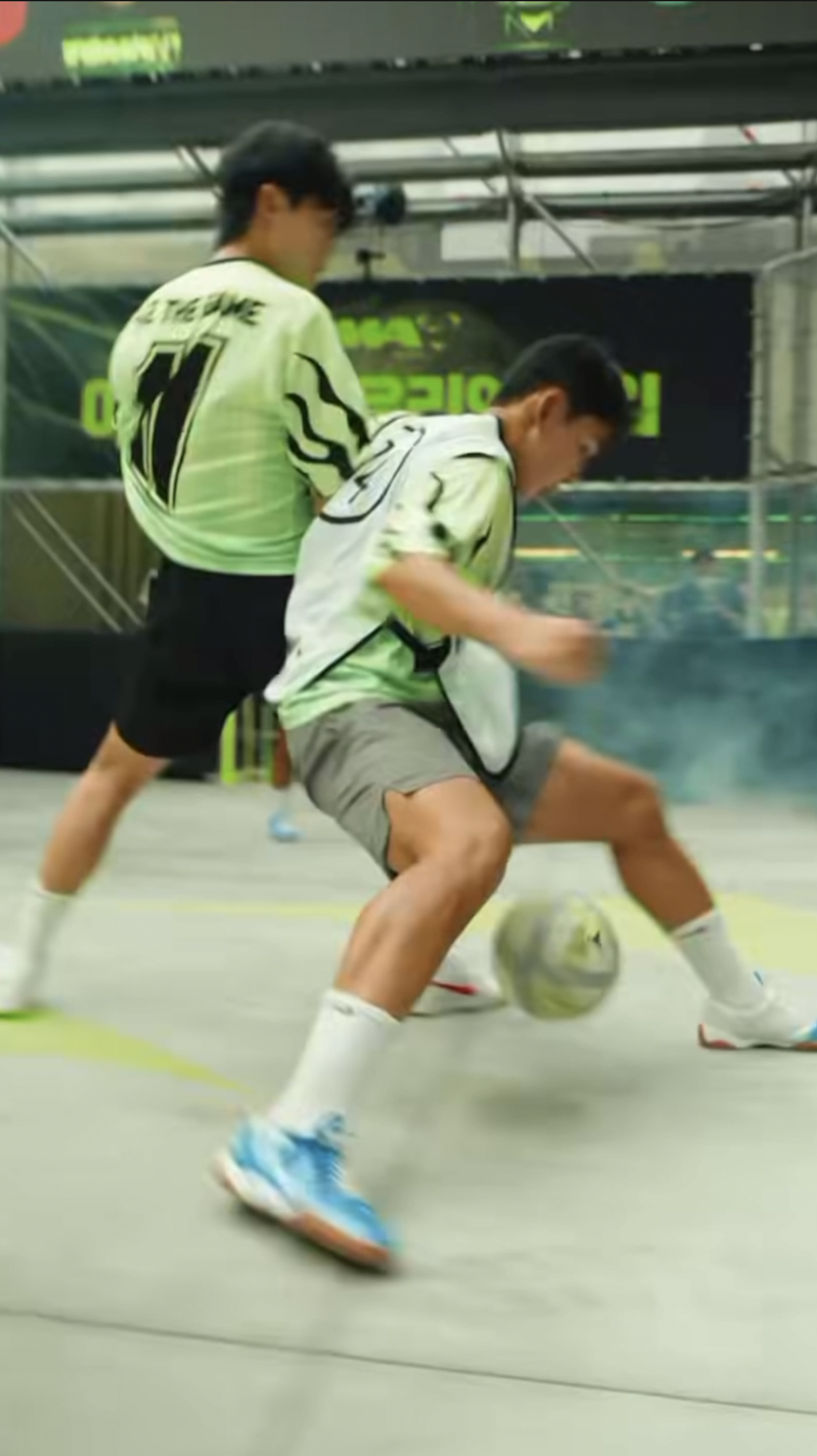 Two young boys wearing sports jerseys and shorts playing indoor soccer, competing for the soccer ball.