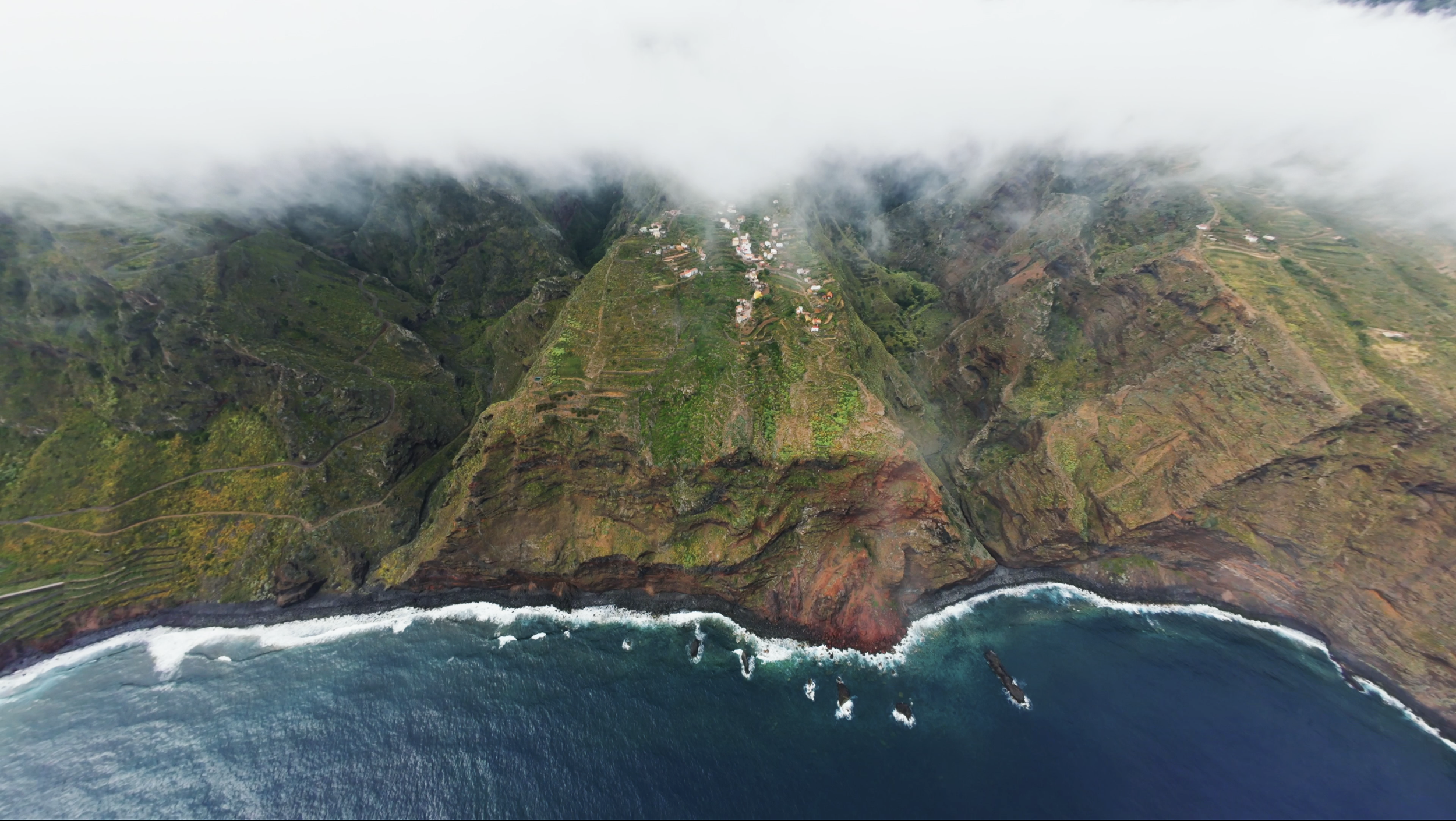 Aerial view of lush green cliffs and coastline with waves crashing on the shore, partially covered by fog.