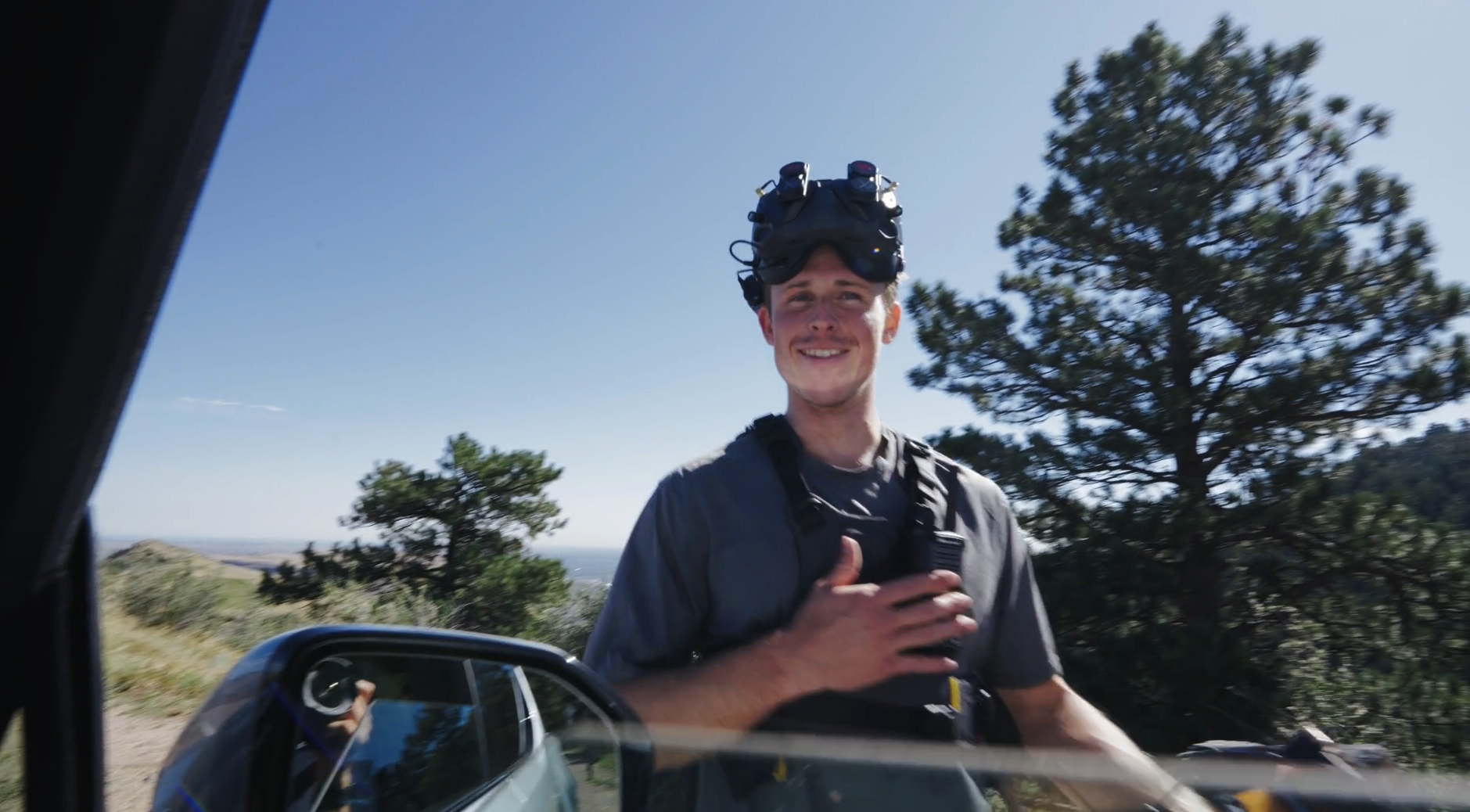 A smiling young man wearing a helmet with mounted gear and a gray T-shirt, standing outdoors beside a vehicle with a side mirror, in a landscape with trees and a clear blue sky.