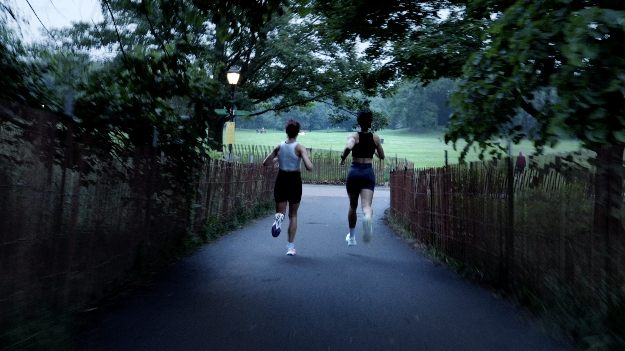 Two women running on a paved path through a park, surrounded by trees and green grass, with a park lamp post and a fence.