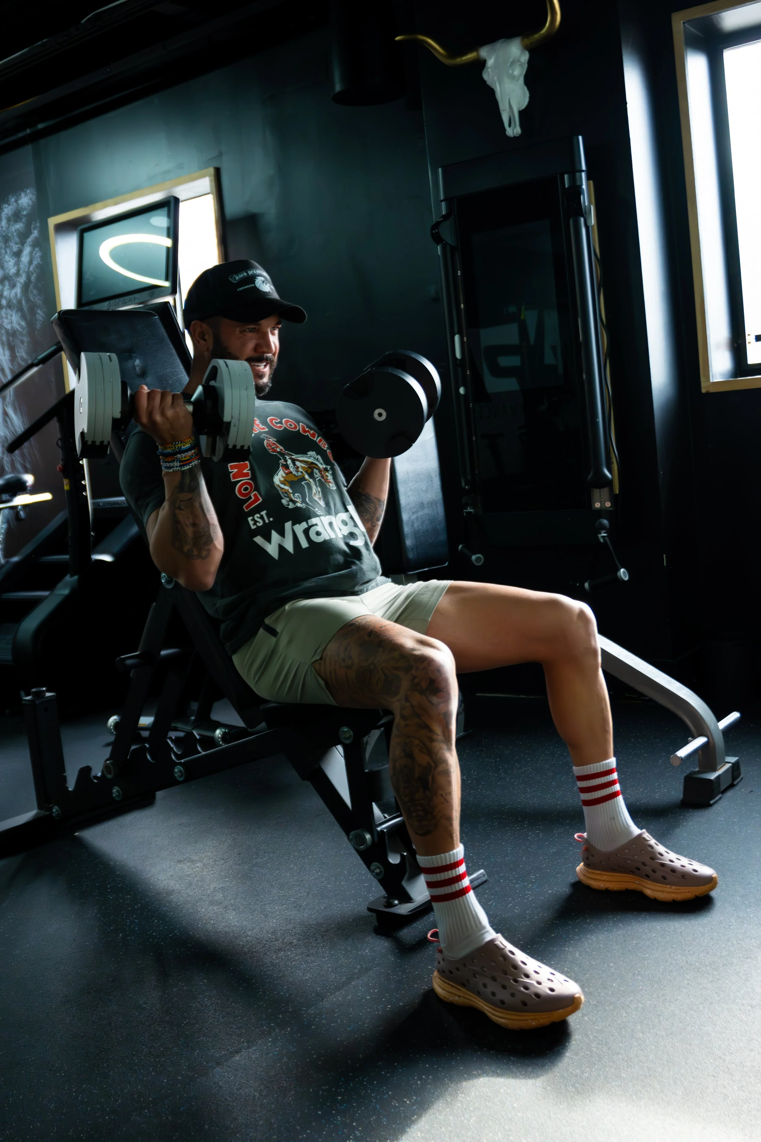 Man in shorts and a t-shirt lifting dumbbells on a seated incline bench in a gym with black walls and windows.
