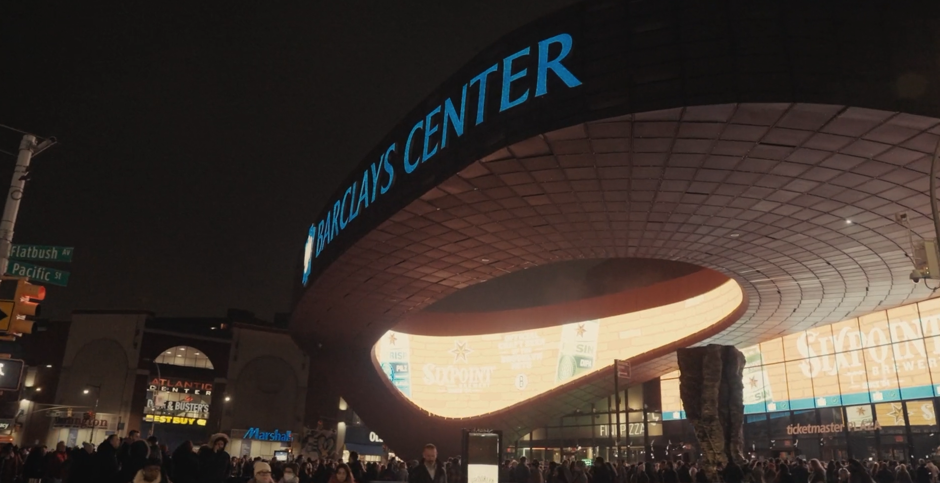 Nighttime scene of the Barclays Center with a crowd of people outside. The distinctive curved roof of the arena is illuminated, displaying the Barclays Center sign in bright blue letters. Surrounding signs for stores and street names are visible, including Flatbush Avenue and Pacific Street.