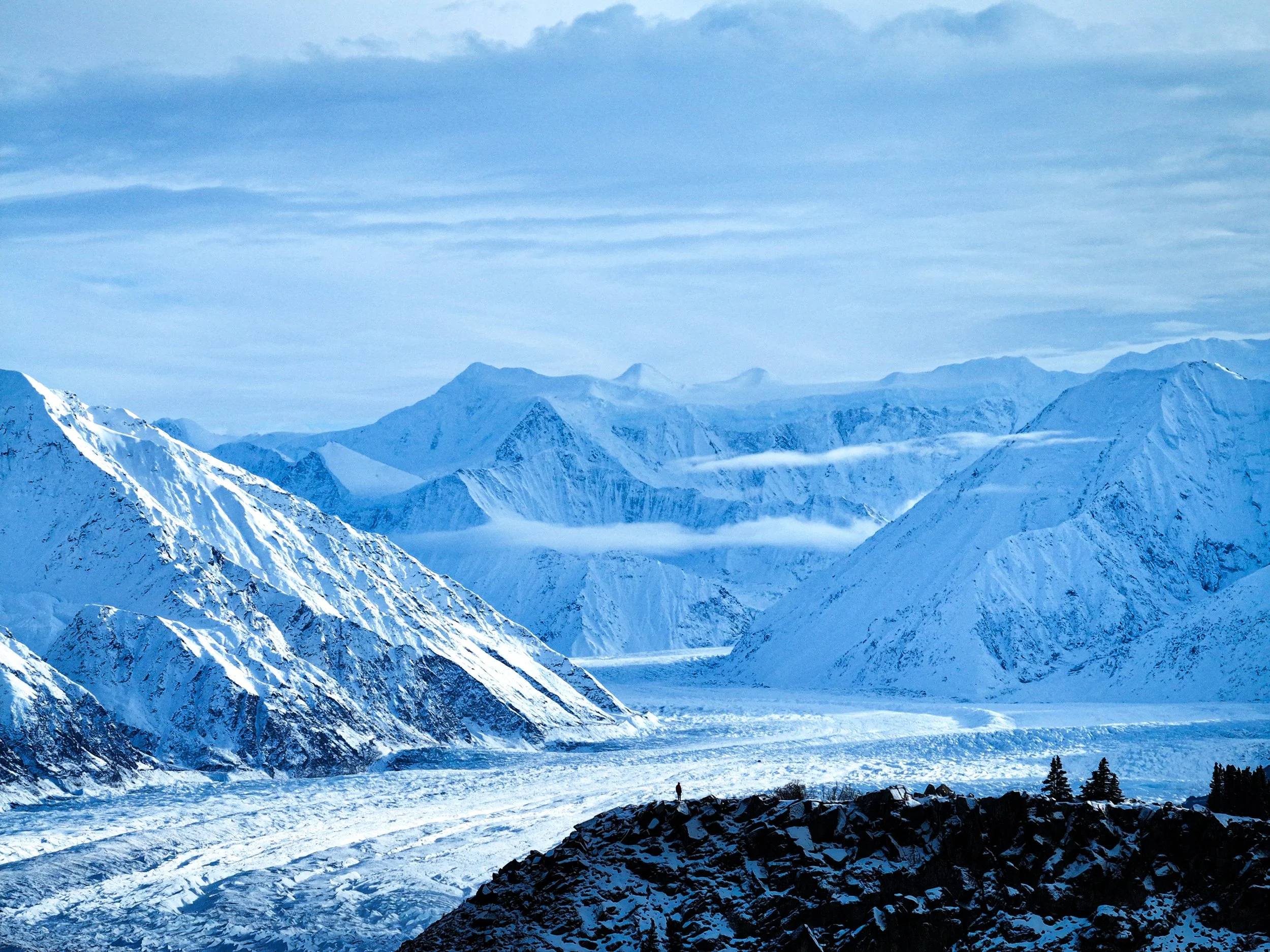 Snow-covered mountain range with glacier in the foreground and a cloudy sky overhead.