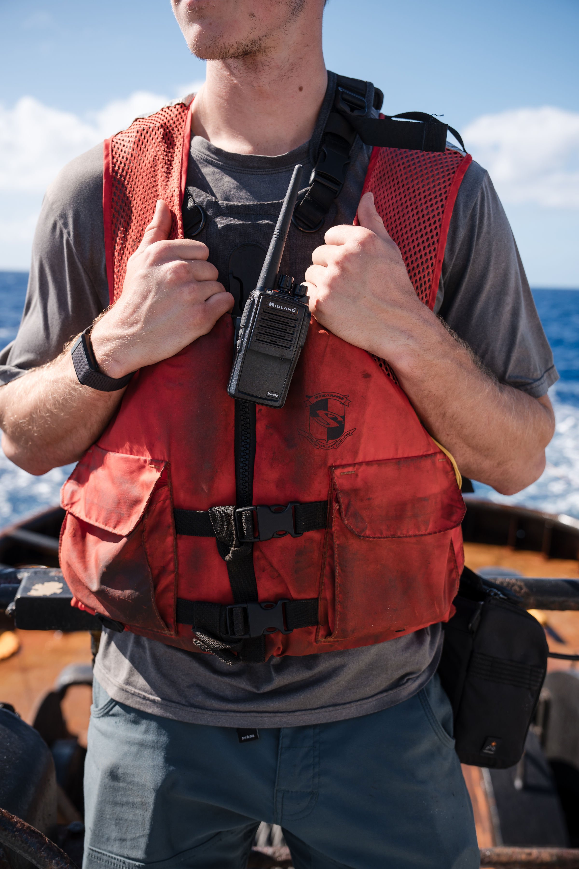 A person on a boat wearing a red life jacket, holding a walkie-talkie, with the ocean in the background.