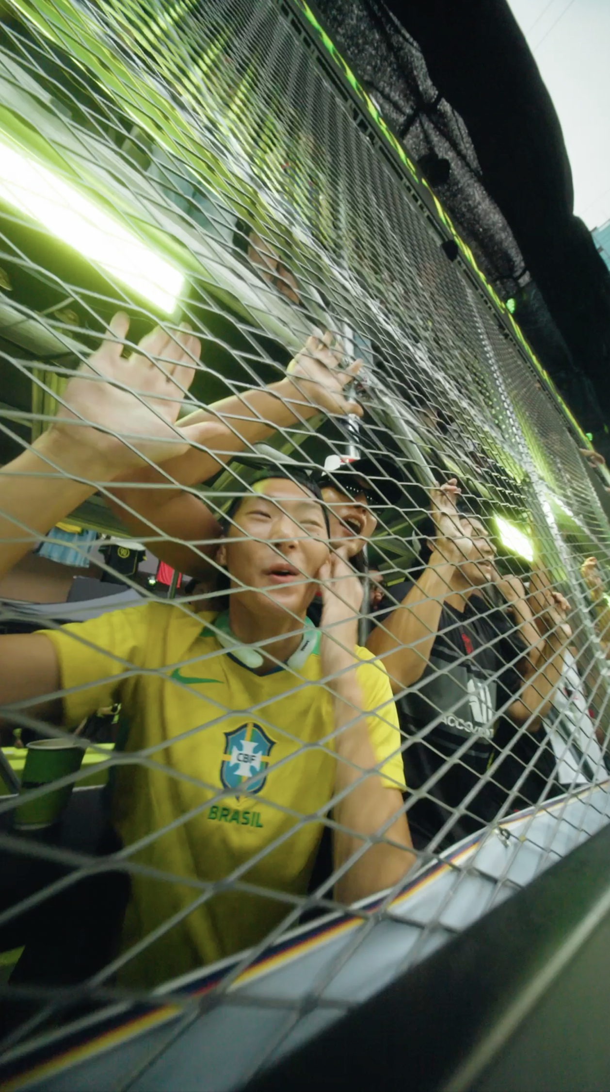 People behind a fencing net, cheering and celebrating at a sporting event, with one person wearing a Brazil national football team shirt.