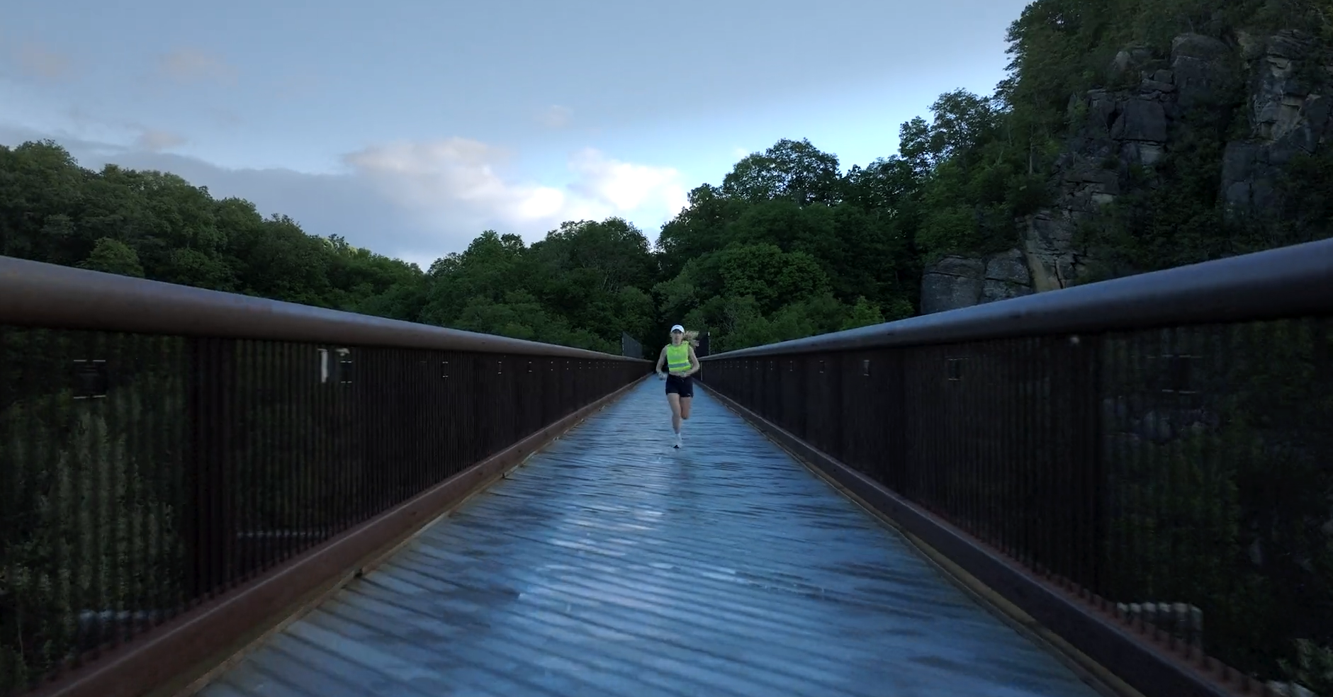 A person running on a long, narrow suspension bridge surrounded by green trees and rocky hills.