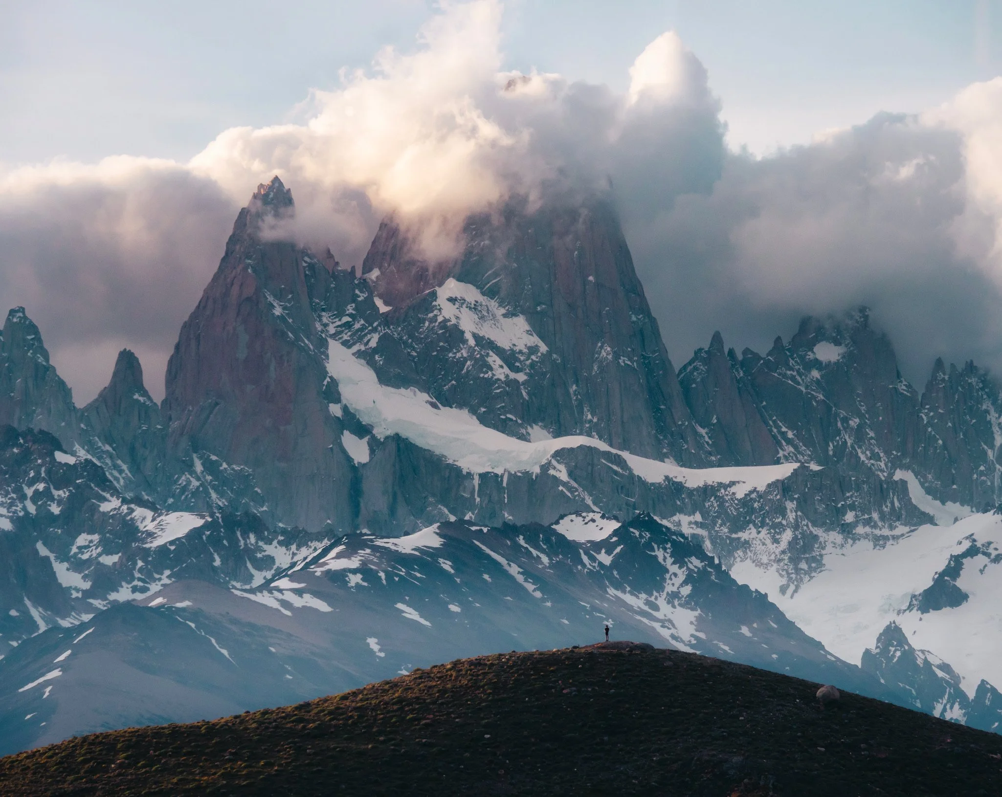 Snow-capped mountain peaks partially shrouded by clouds with a person standing on a grassy hill in the foreground.