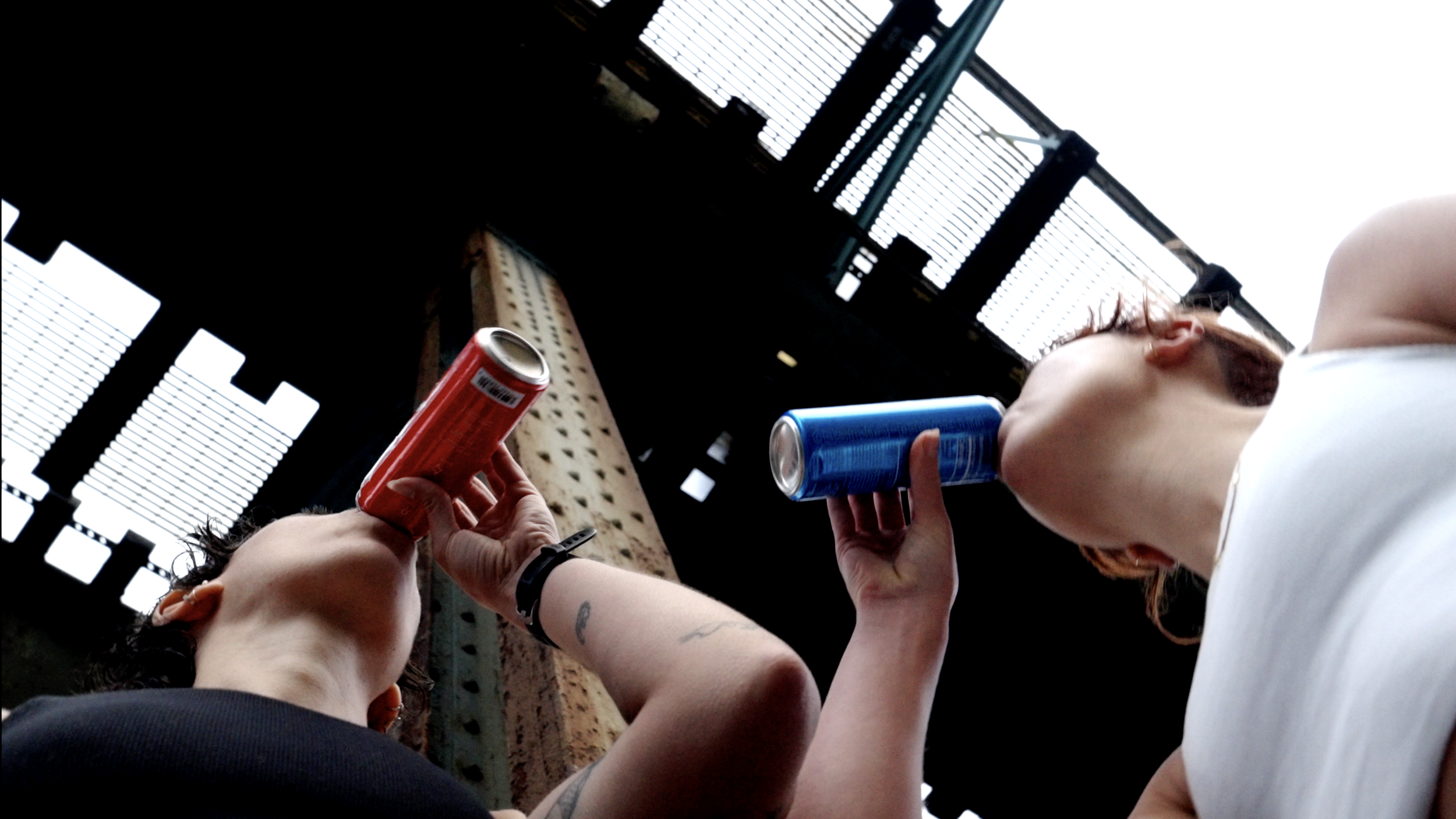 Two women are sitting outdoors, each holding a can of soda, with an industrial background visible above them.