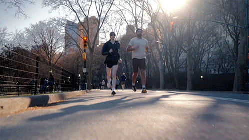 Two people jogging on a city street during sunrise with leafless trees and buildings in the background.