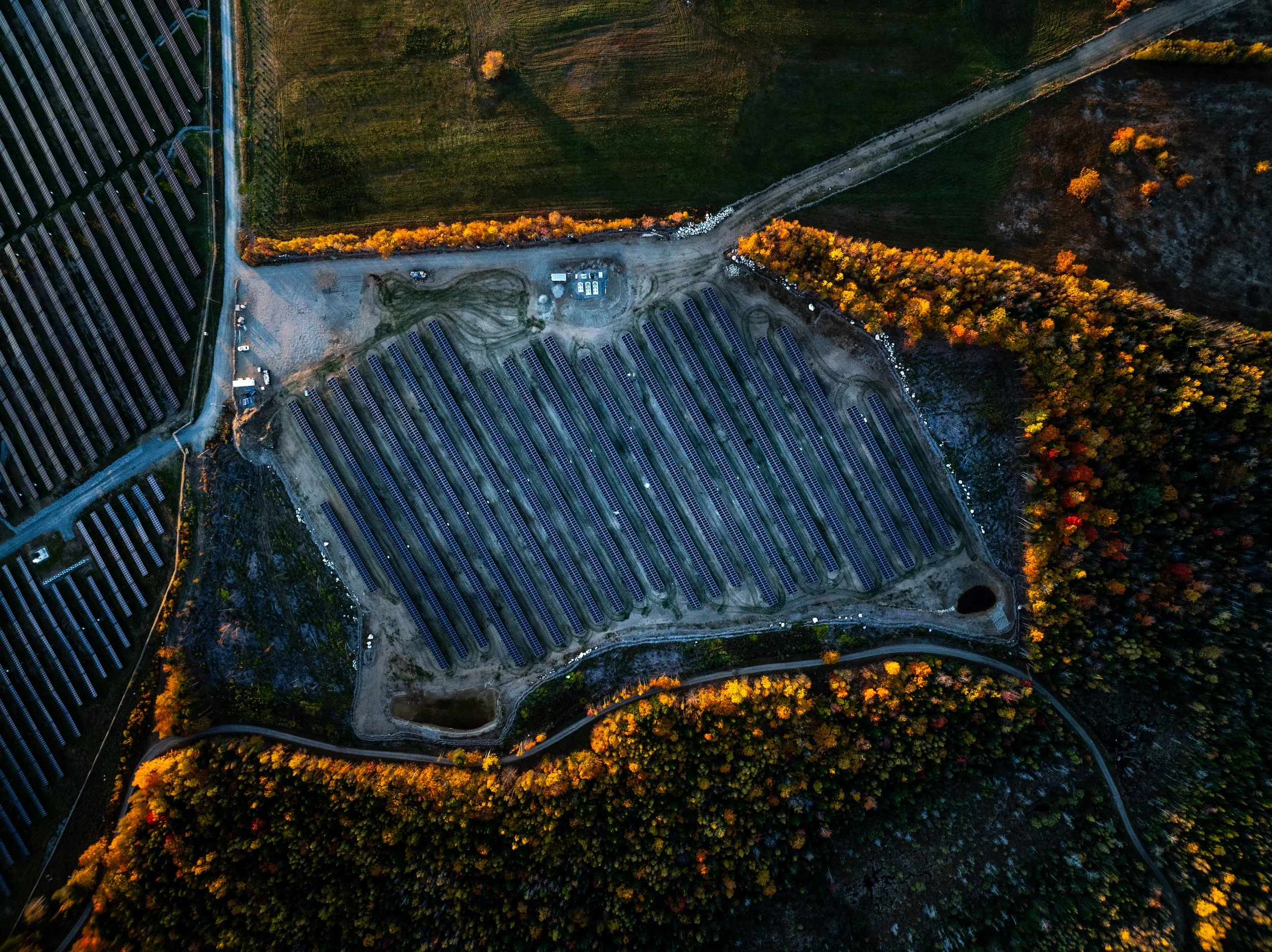 Aerial view of a solar farm with rows of solar panels surrounded by trees displaying fall colors, located in a rural area with dirt roads.