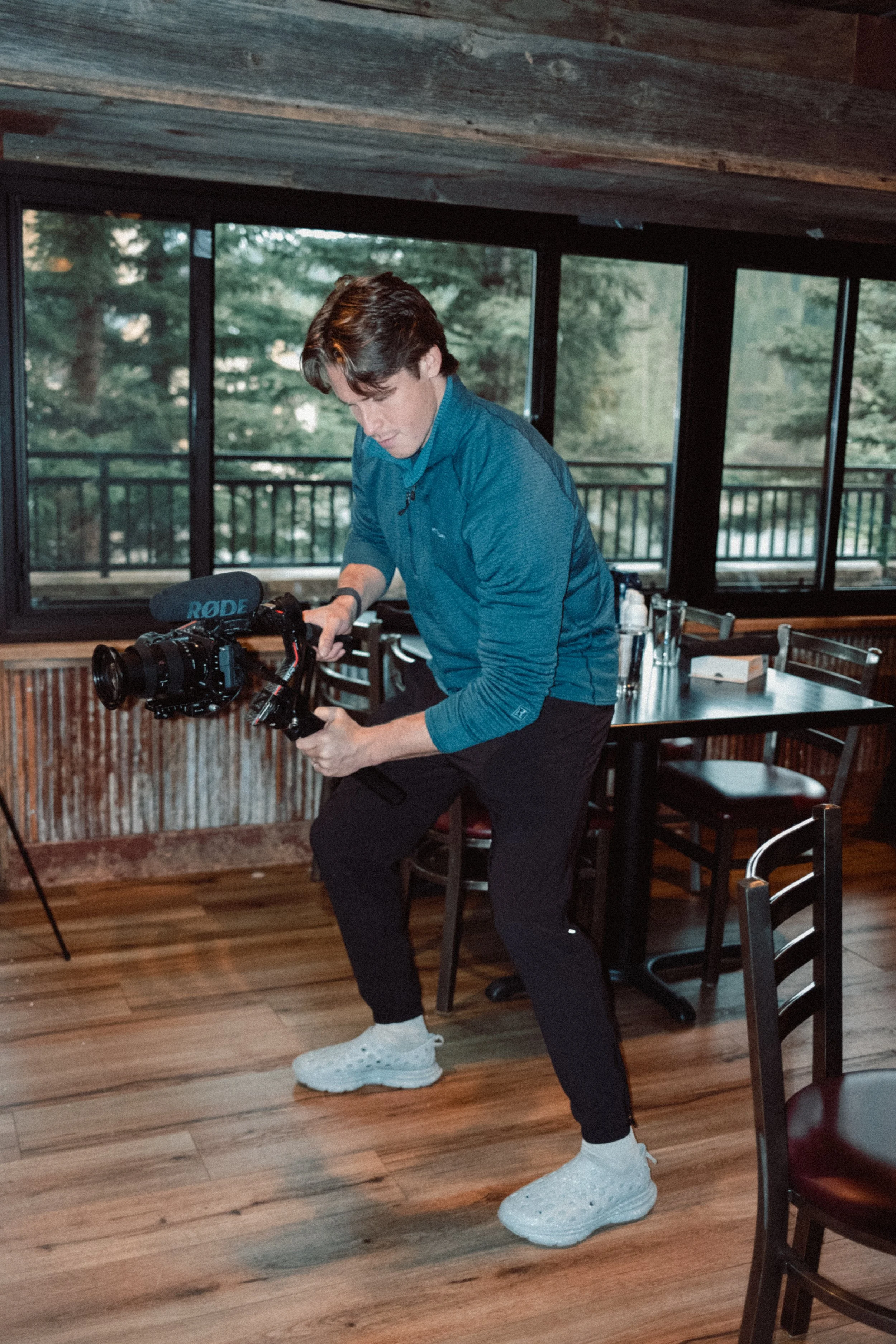 A person wearing a blue jacket and black pants is adjusting a camera with a RODE microphone inside a wooden room with large windows showing trees outside.