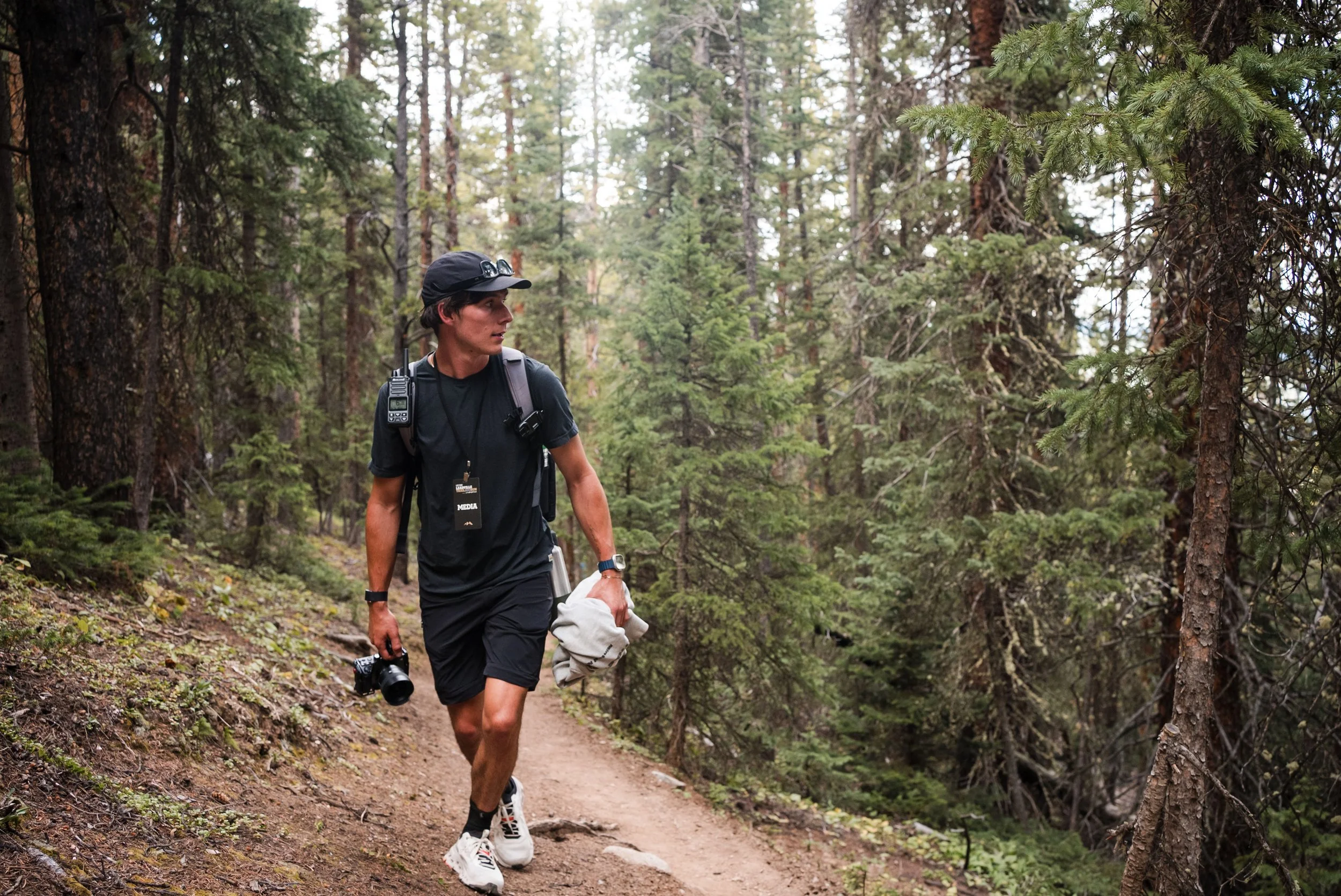 A young man walking on a dirt trail in a dense forest with tall trees, wearing a black t-shirt, shorts, a cap, and carrying a backpack, camera, and cloth.