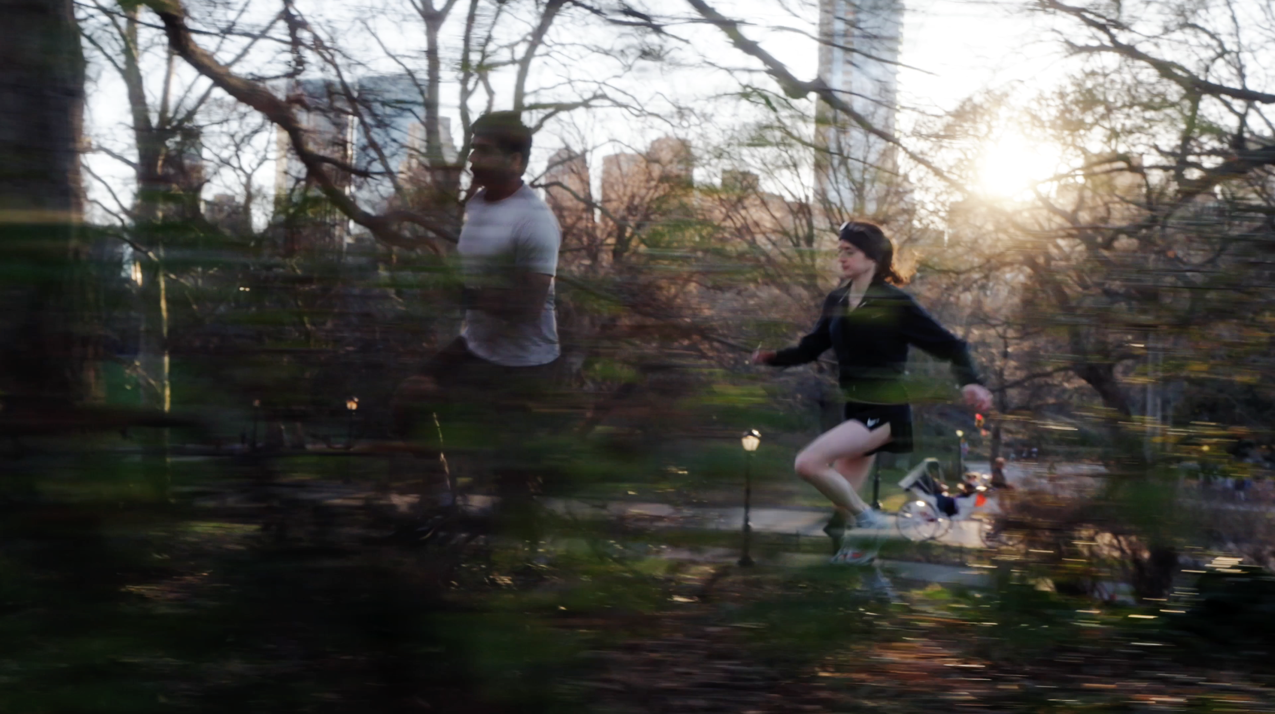 A woman jogging with a backpack and a man jogging nearby in a park at sunset, trees in the background, with some motion blur.