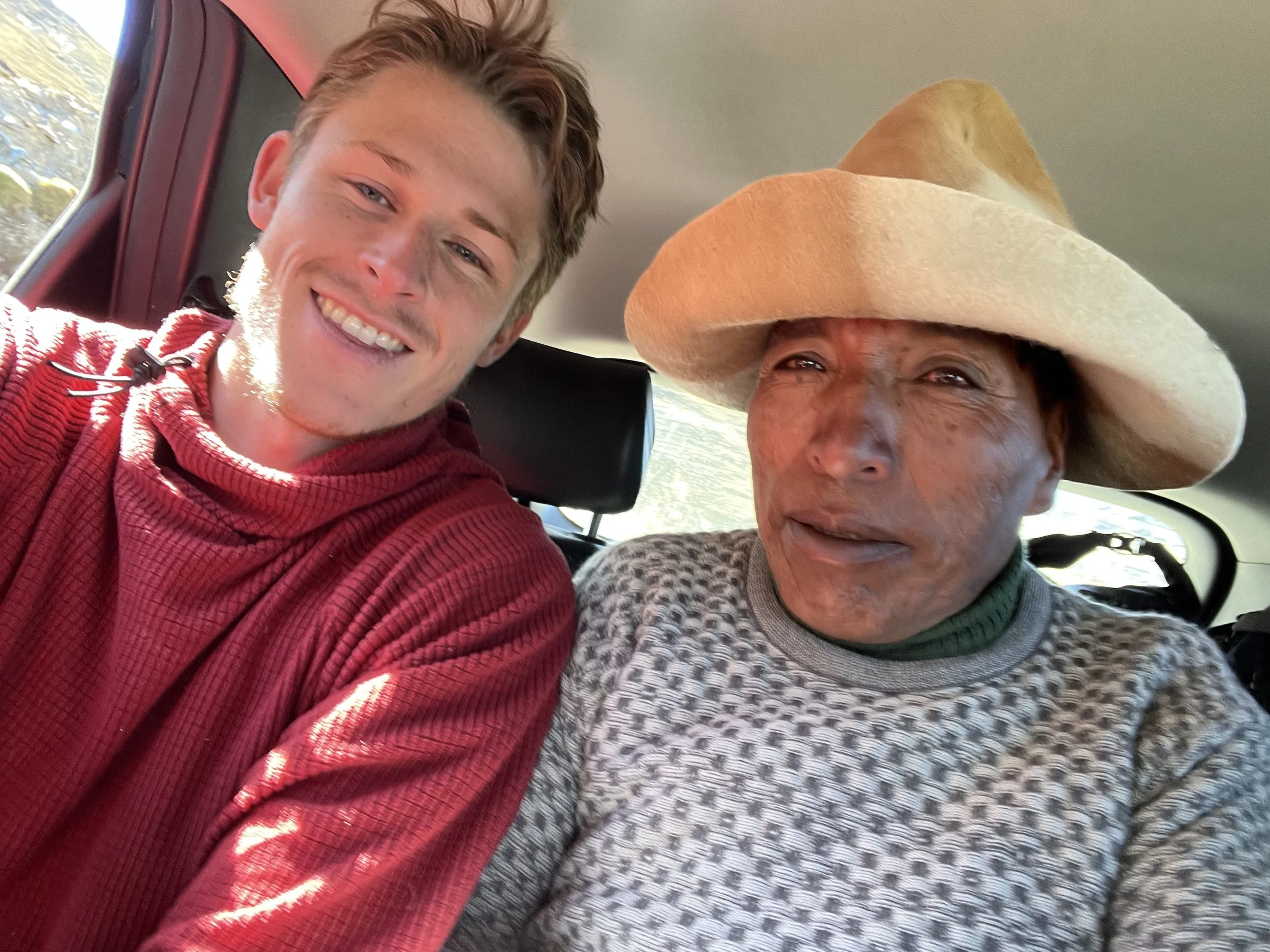 A young man and an elderly woman sitting in a car, smiling for a selfie. The young man is wearing a red sweater, and the elderly woman is wearing a large hat and a patterned sweater.