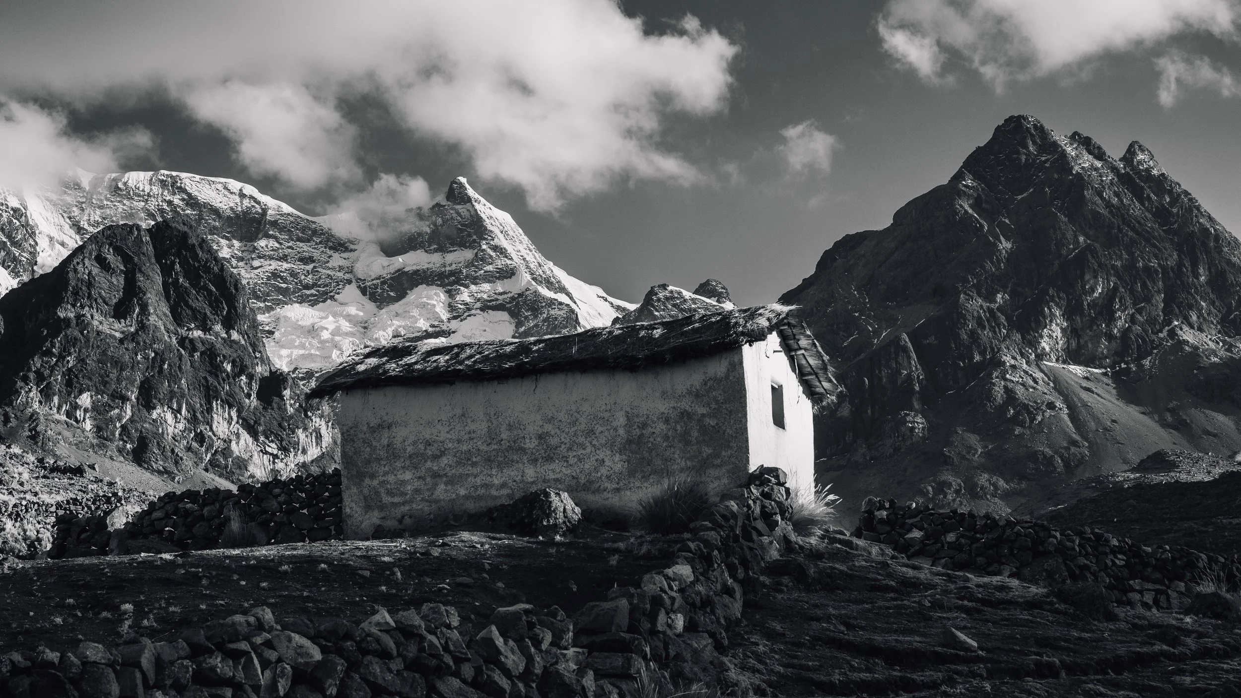 A small white house with a sloped roof in a mountainous landscape with snow-capped peaks and clouds in the background.