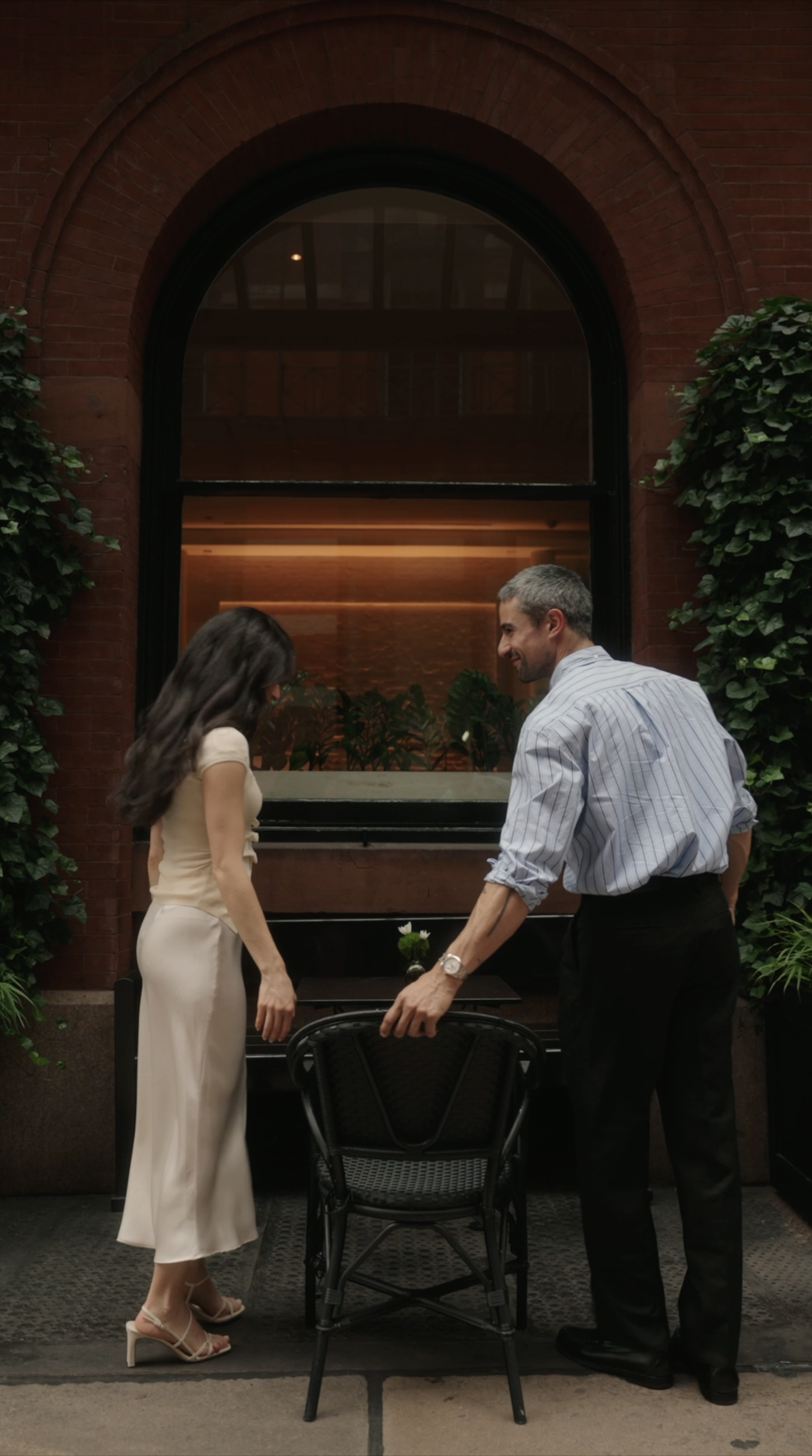 A man and woman standing outside near a black table and chair, with a brick building and large window in the background, engaging in an interaction with a small bouquet of flowers on the table.