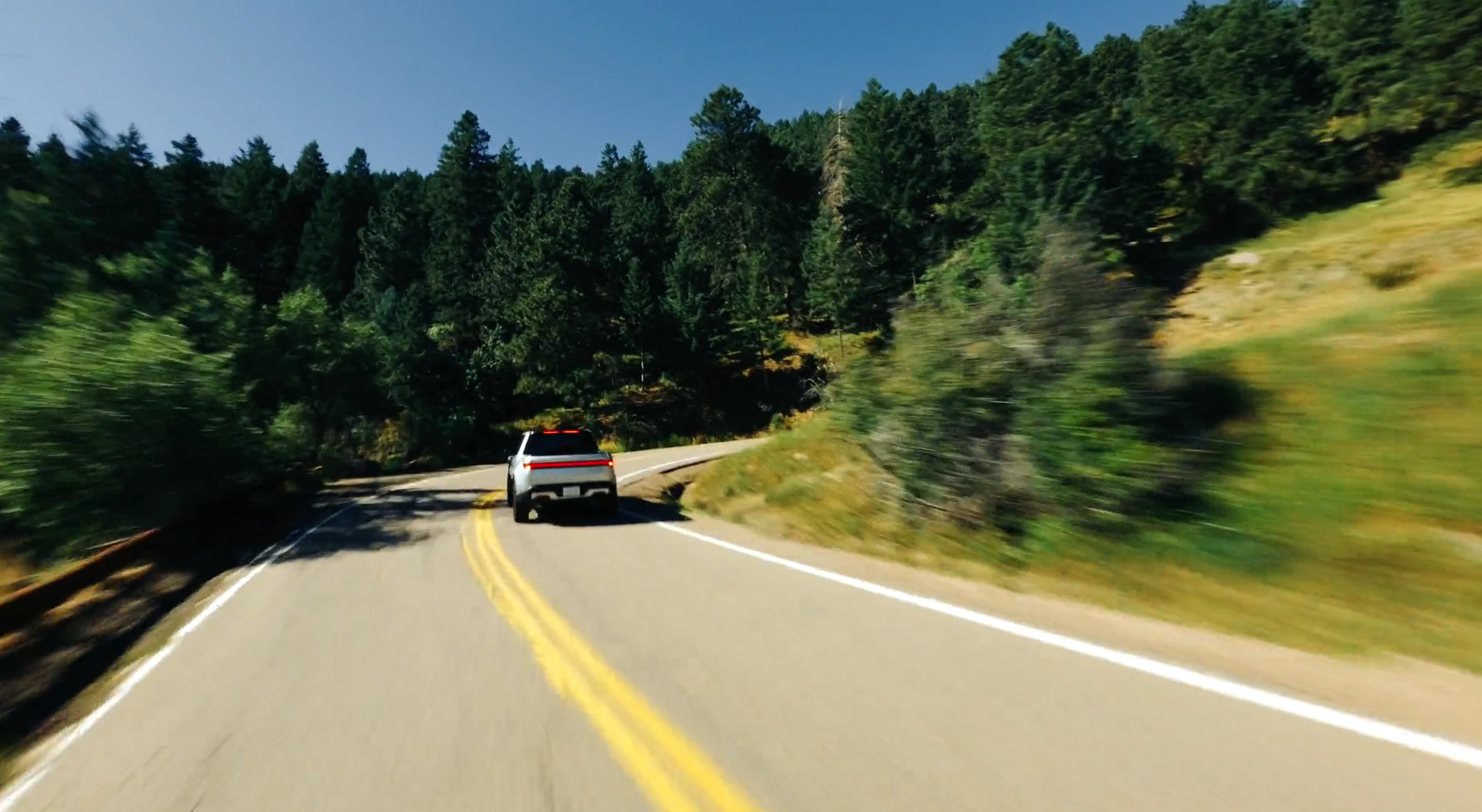 A white vehicle driving on a winding mountain road surrounded by trees and greenery under a clear blue sky.