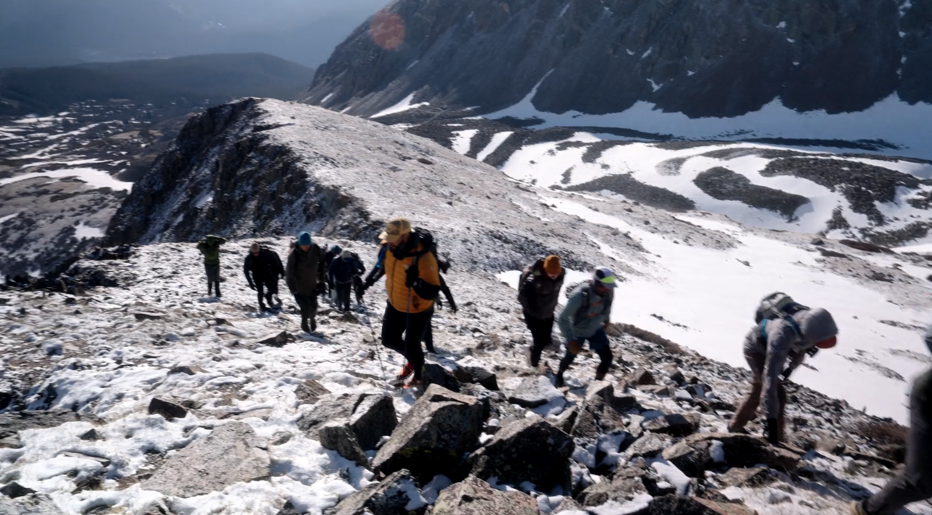 Group of hikers climbing a snow-covered mountain trail with rocky terrain, snow patches, and mountain peaks in the background.