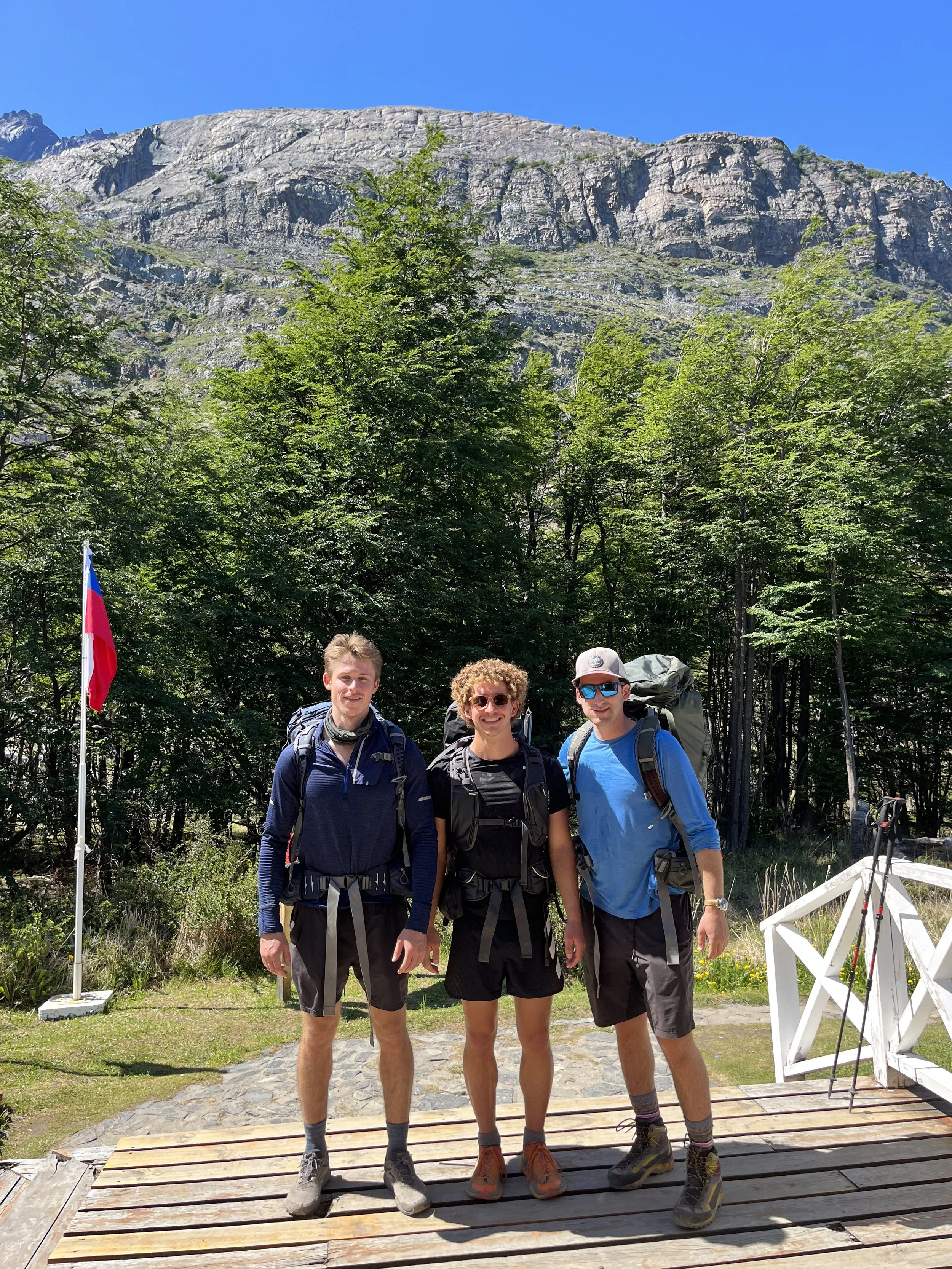 Three young men with backpacks and hiking gear standing on a wooden platform, smiling. Behind them are trees, a mountain in the background, and a flag on a pole, with a clear blue sky overhead.