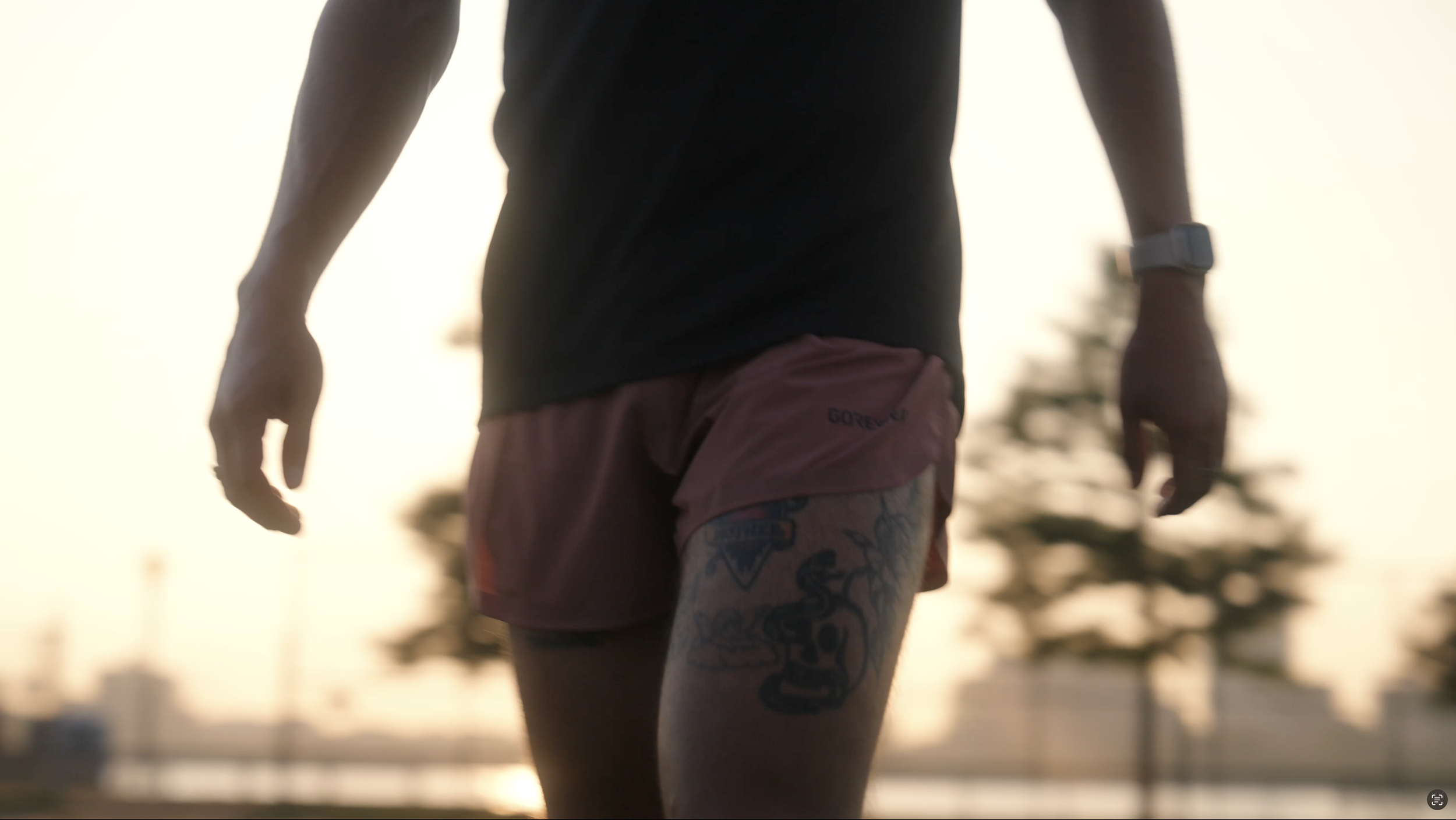 A person wearing athletic clothing and running shorts, with a tattoo on their thigh, is captured from the waist down outdoors during sunset.