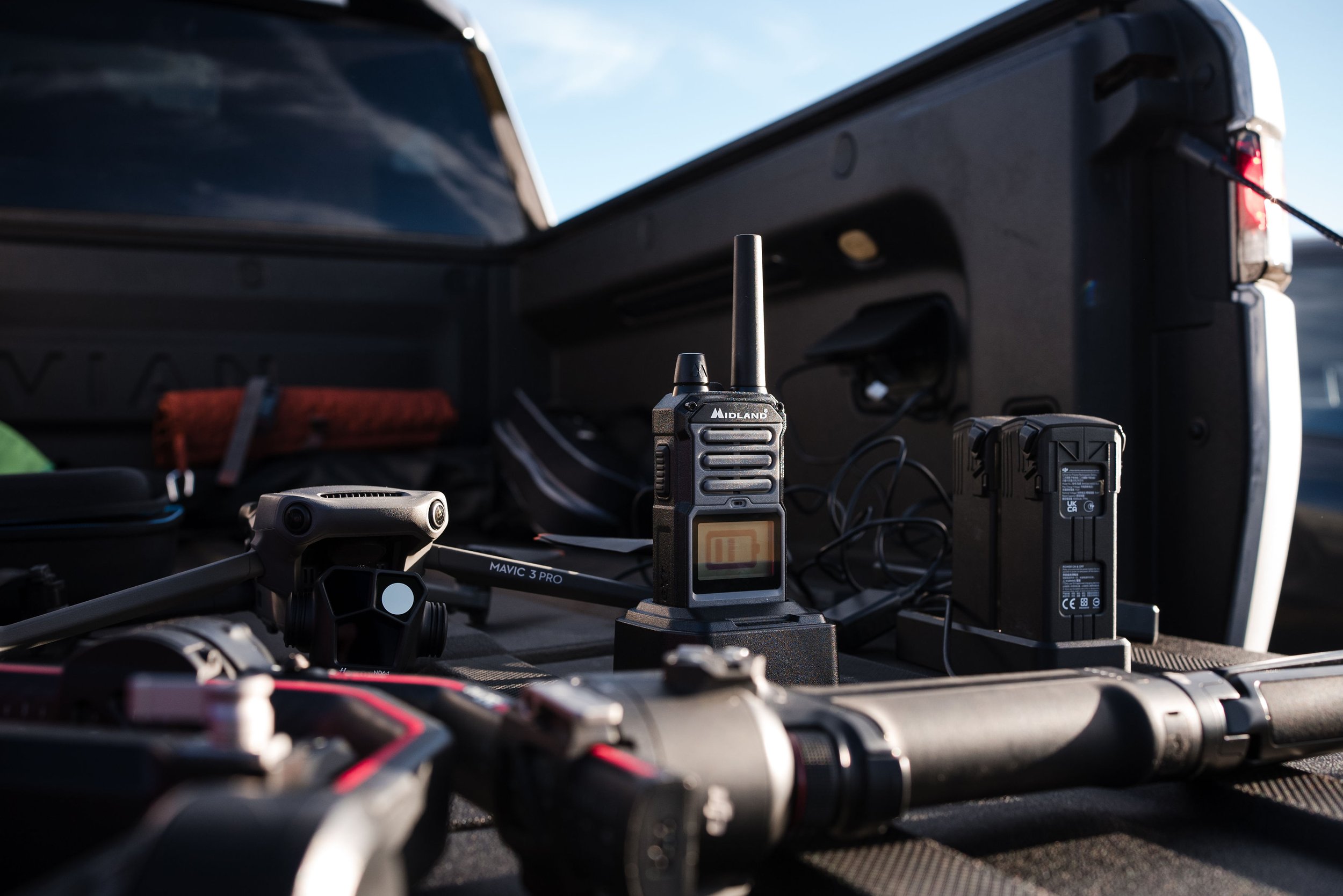 A drone, a walkie-talkie, and camera batteries inside a vehicle's hatchback with outdoor scenery in the background.