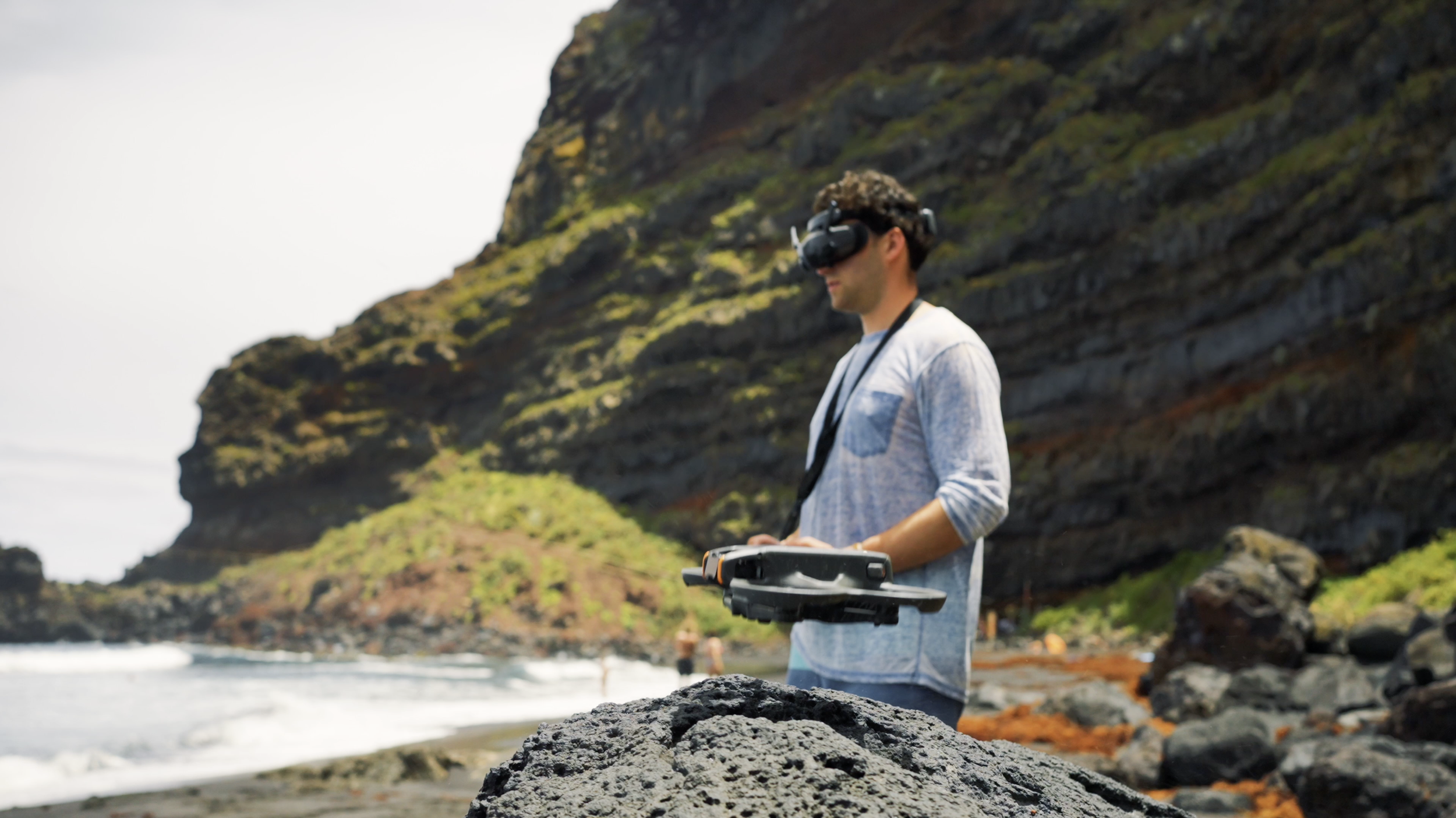 A man wearing a VR headset and holding a drone controller on a rocky beach with cliffs and ocean waves in the background.