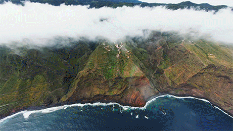 Aerial view of green mountain cliffs along a coastline with fog and ocean waves.