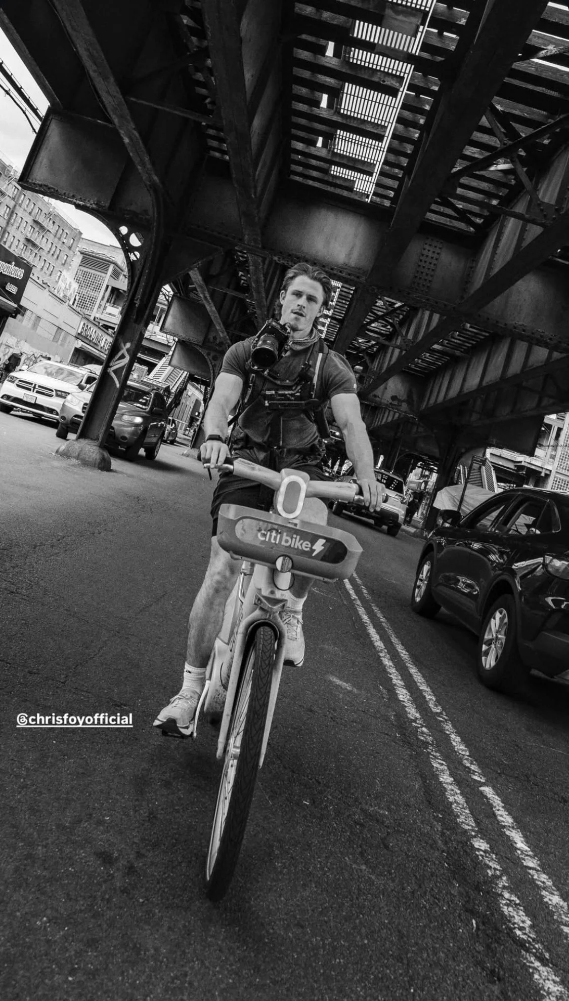 A man riding a Citi Bike on an urban street under an elevated train track. He is carrying a camera around his neck and has a backpack.