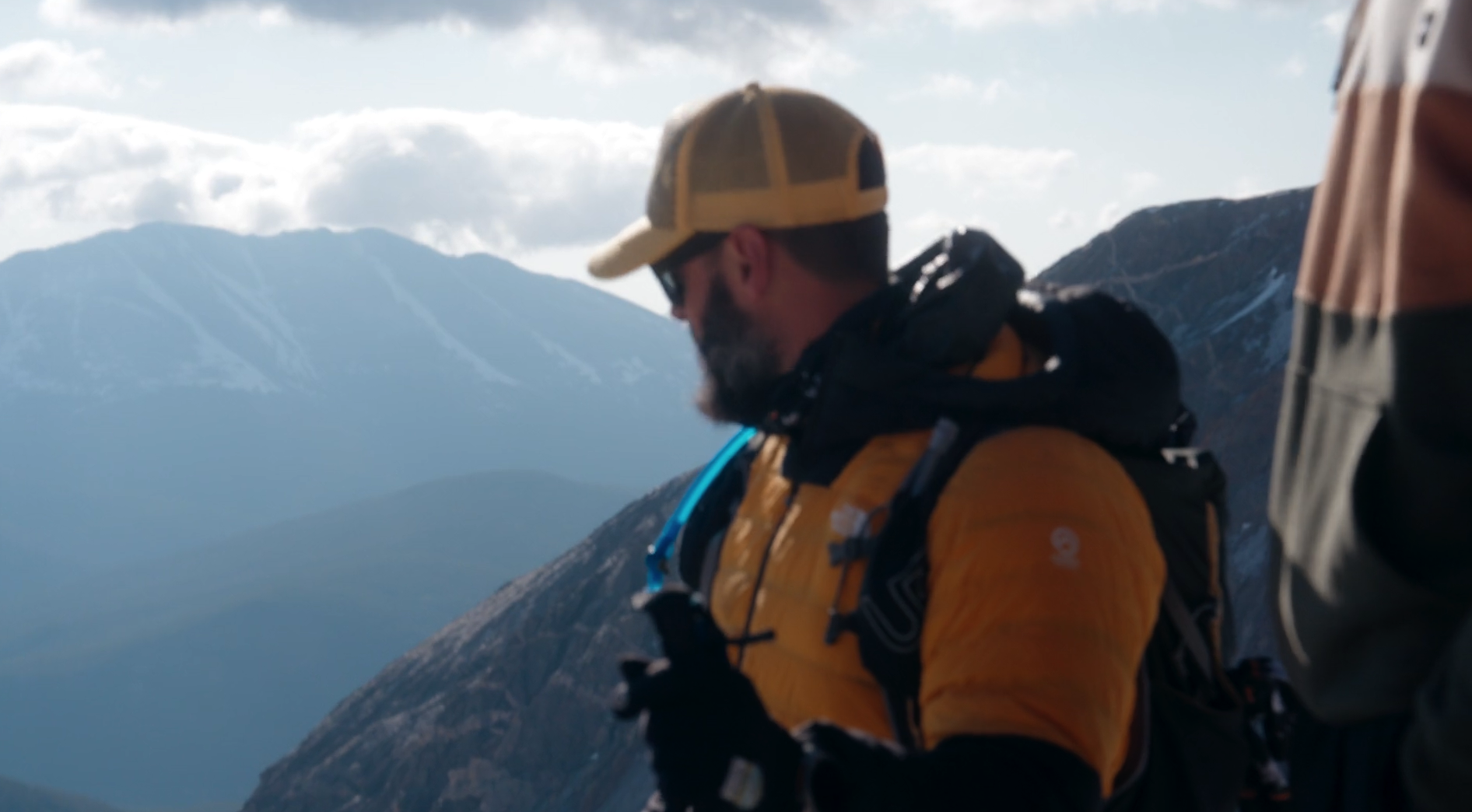A man with a beard wearing a yellow cap, yellow jacket, sunglasses, and a backpack, standing outdoors in a mountainous area with snow-capped peaks in the background.